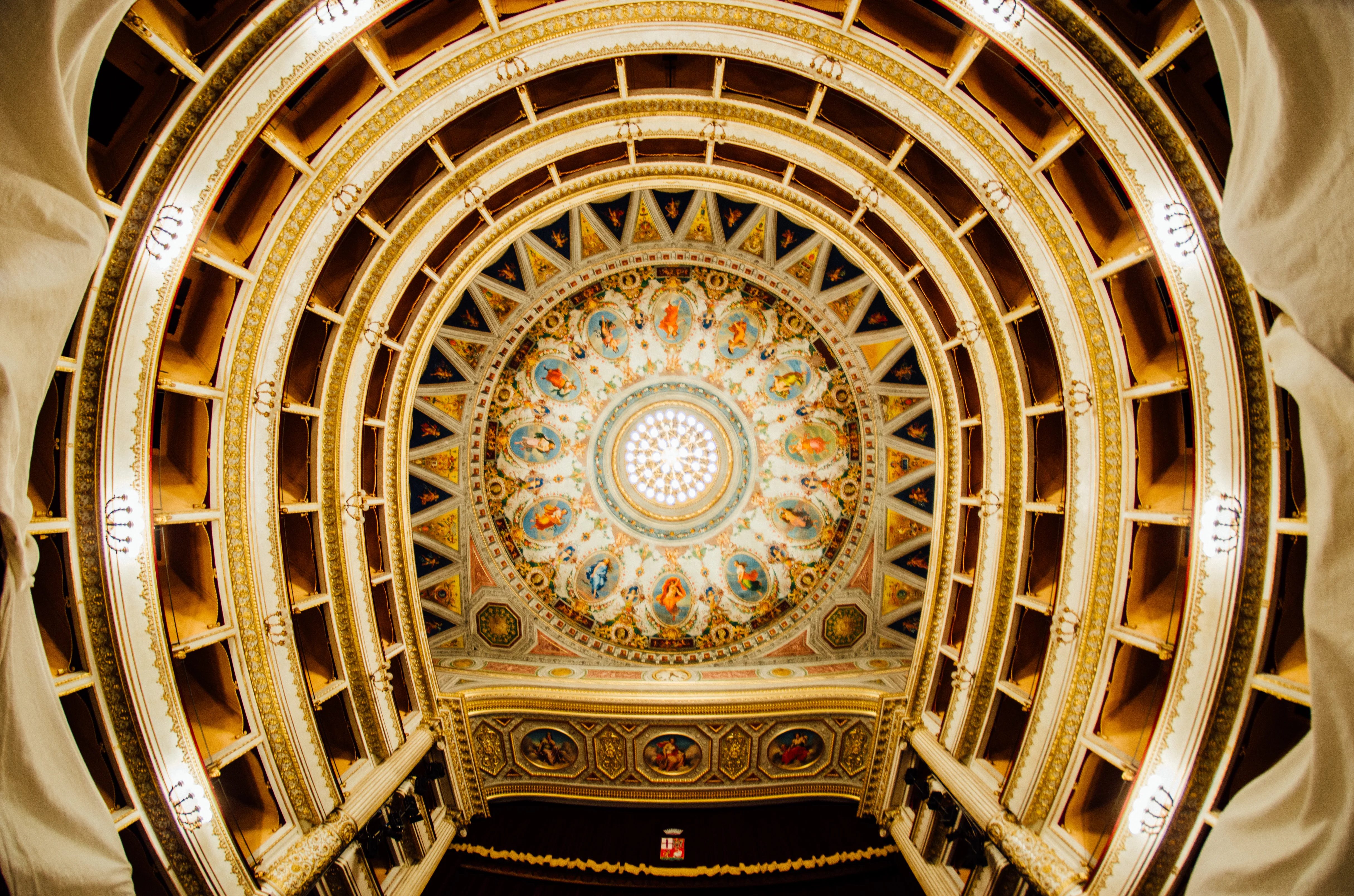 Vue en contre-plongée du plafond d'un opéra classique orné de fresques colorées, d'une coupole centrale avec oculus lumineux et de plusieurs niveaux de balcons dorés en fer à cheval.