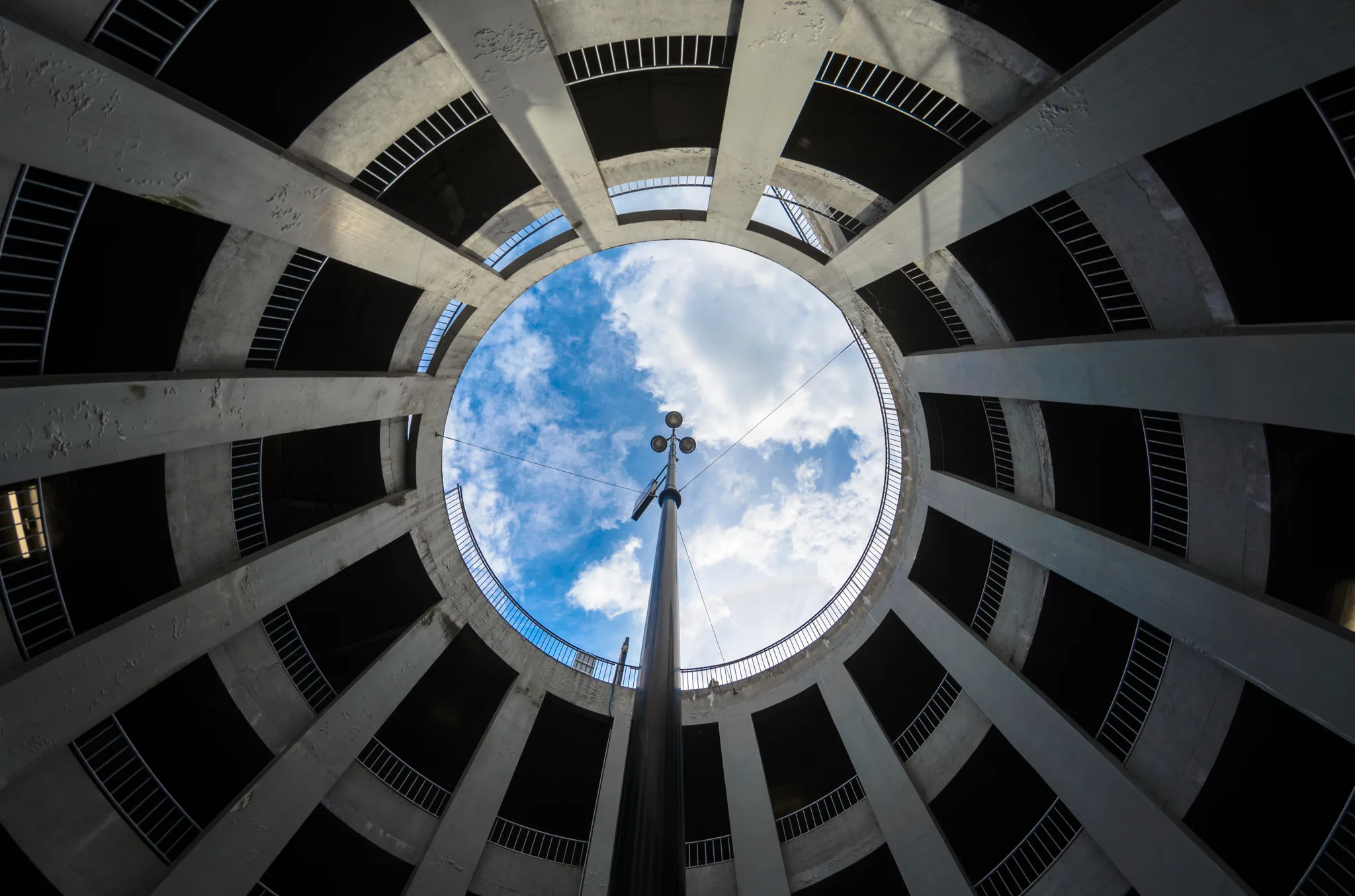 Vue en contre-plongée depuis le bas d'un parking circulaire en béton, avec des piliers rayonnants encadrant une ouverture ronde vers un ciel bleu nuageux, traversée par un mât d'éclairage central.