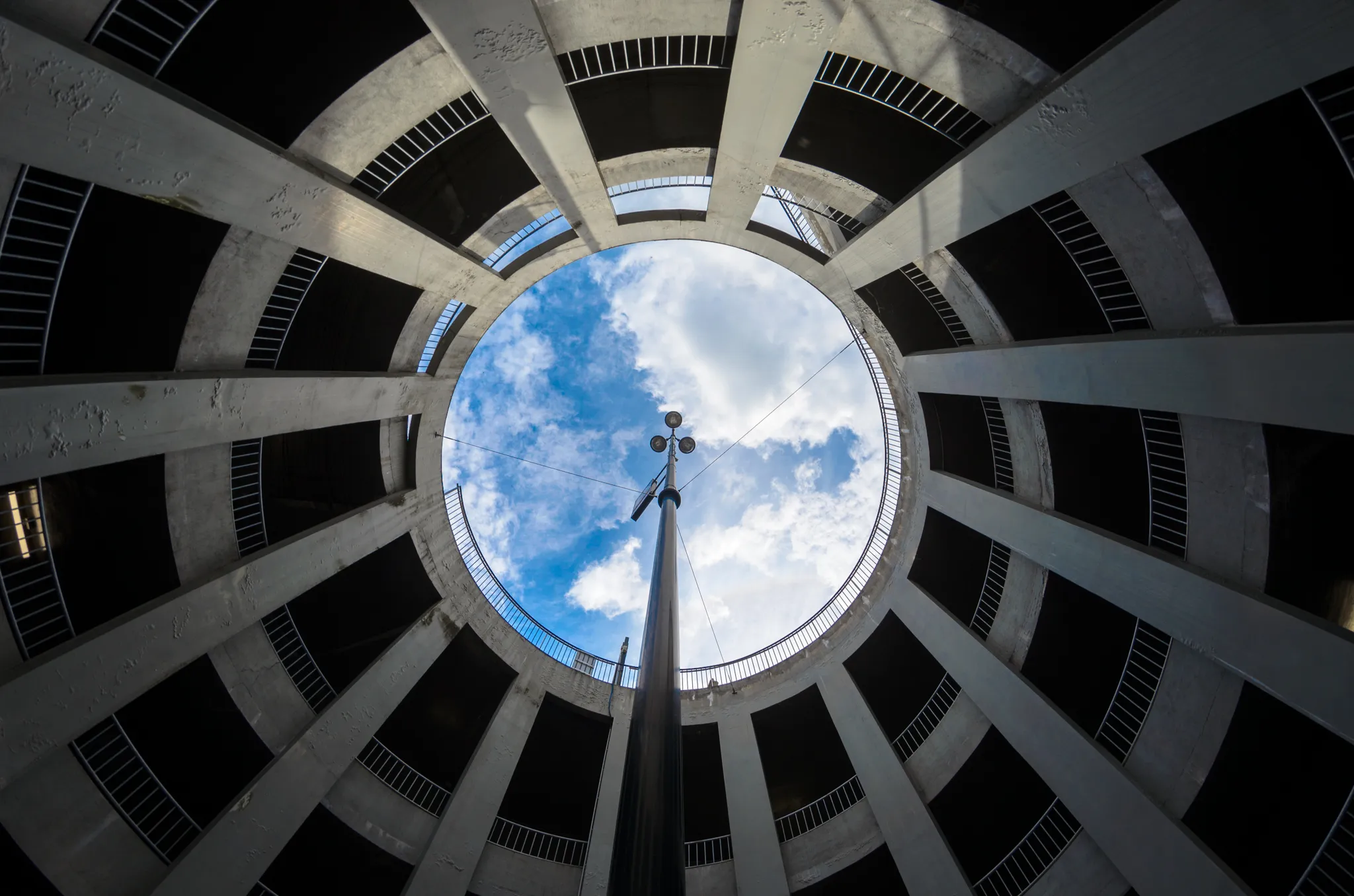 Vue en contre-plongée depuis le bas d'un parking circulaire en béton, avec des piliers rayonnants encadrant une ouverture ronde vers un ciel bleu nuageux, traversée par un mât d'éclairage central.