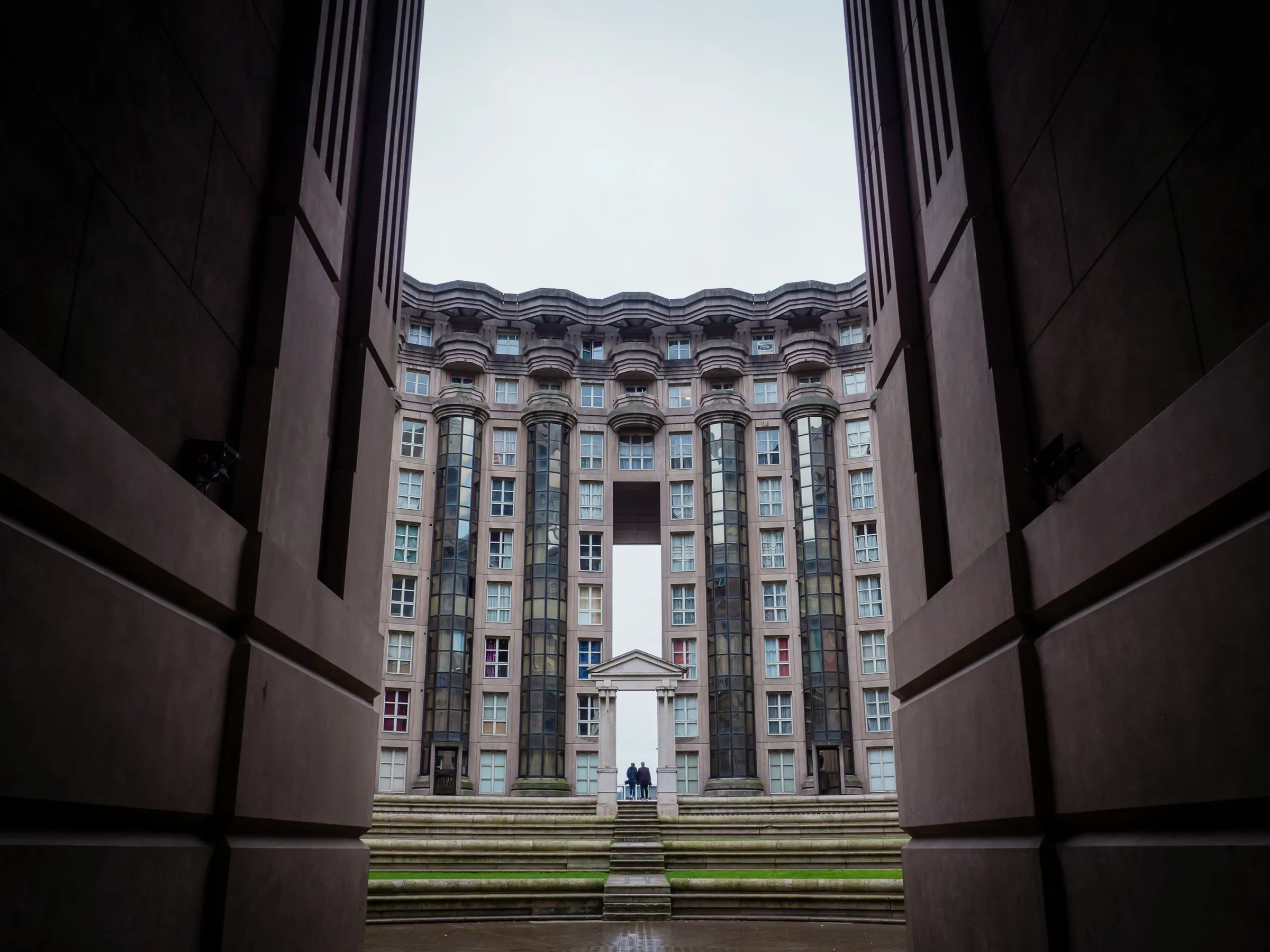Vue en contre-plongée depuis l'intérieur d'un vaste passage voûté donnant sur la cour centrale des Arènes de Picasso, avec sa façade incurvée aux colonnes cylindriques vitrées et un portique néoclassique au fond, sous un ciel gris.