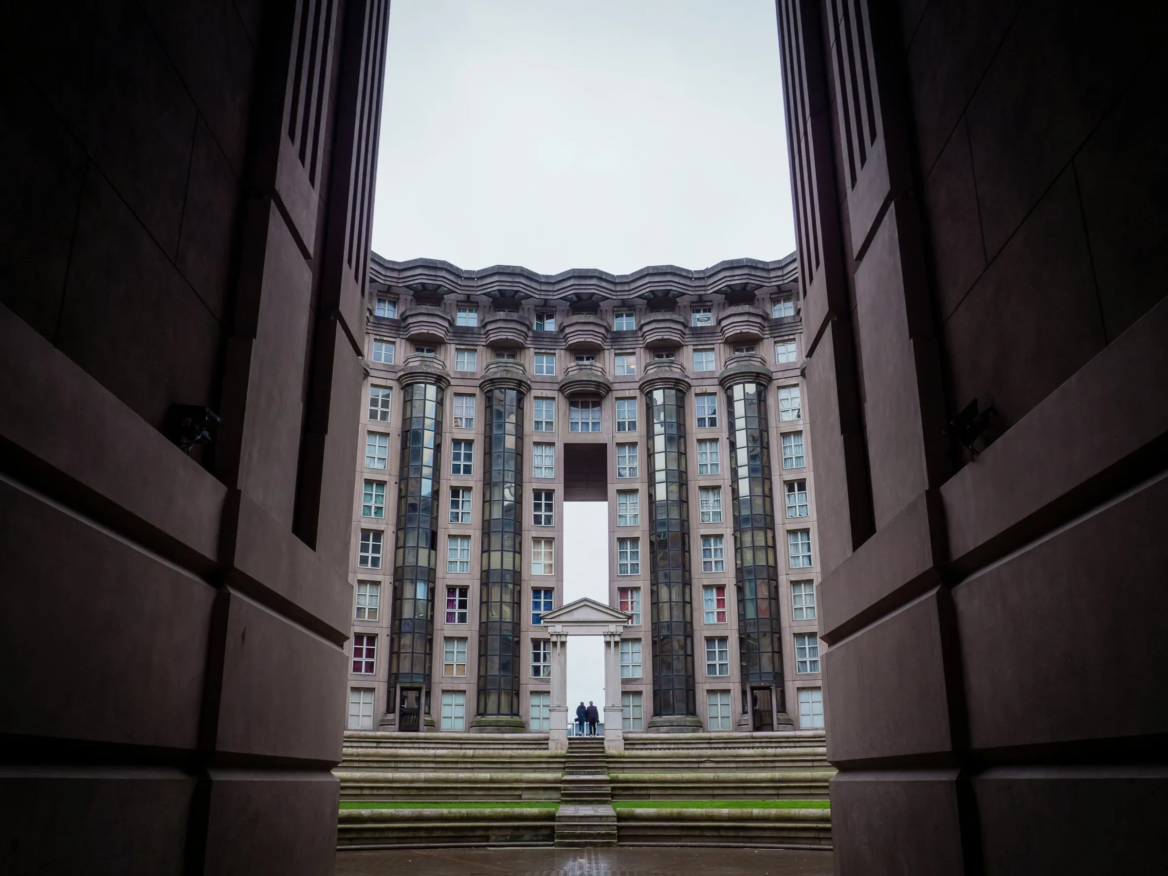 Vue en contre-plongée depuis l'intérieur d'un vaste passage voûté donnant sur la cour centrale des Arènes de Picasso, avec sa façade incurvée aux colonnes cylindriques vitrées et un portique néoclassique au fond, sous un ciel gris.