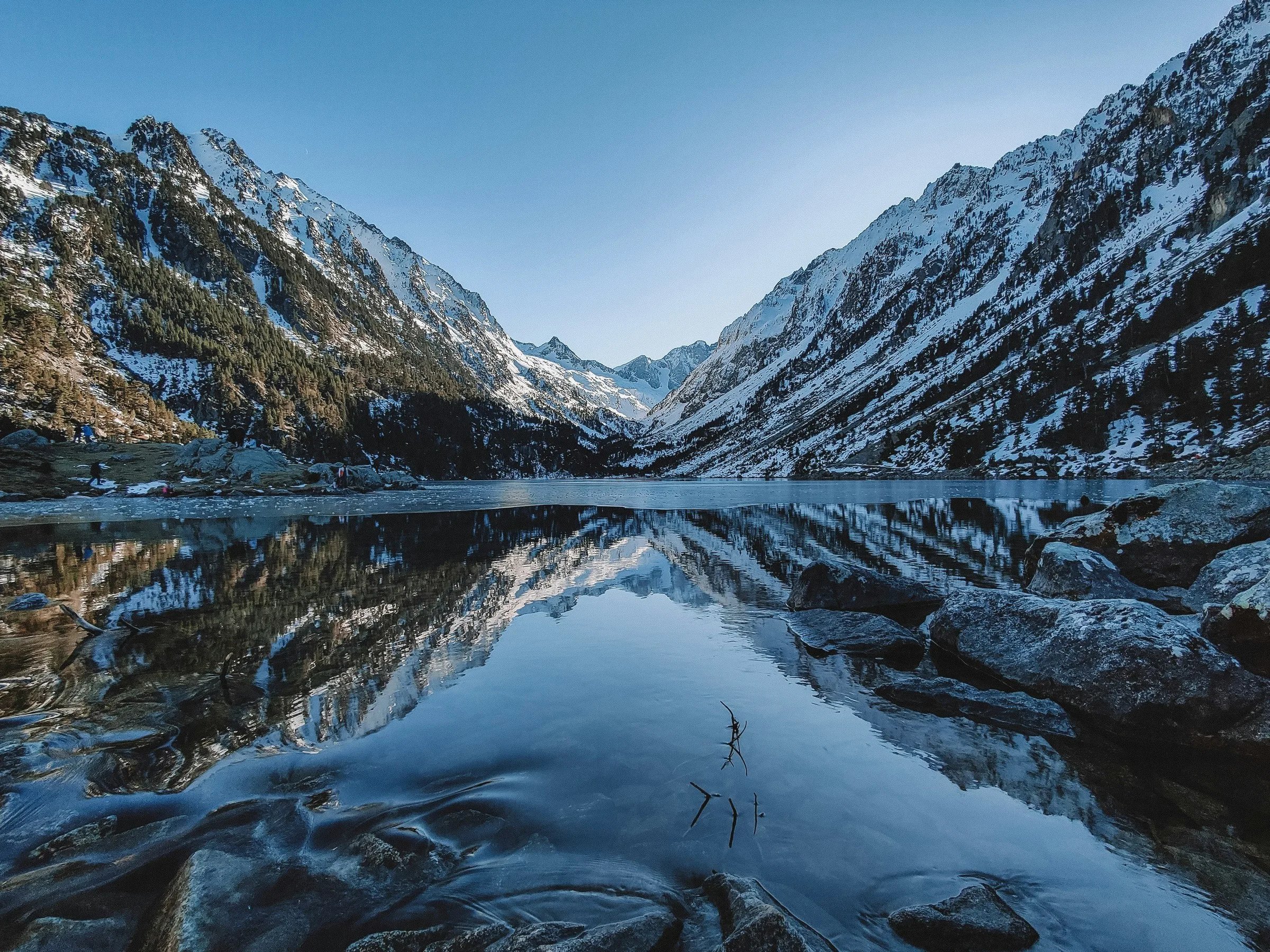 Lac de montagne aux eaux calmes et transparentes reflétant des sommets enneigés encadrés par une vallée glaciaire sous un ciel bleu limpide, avec des rochers au premier plan