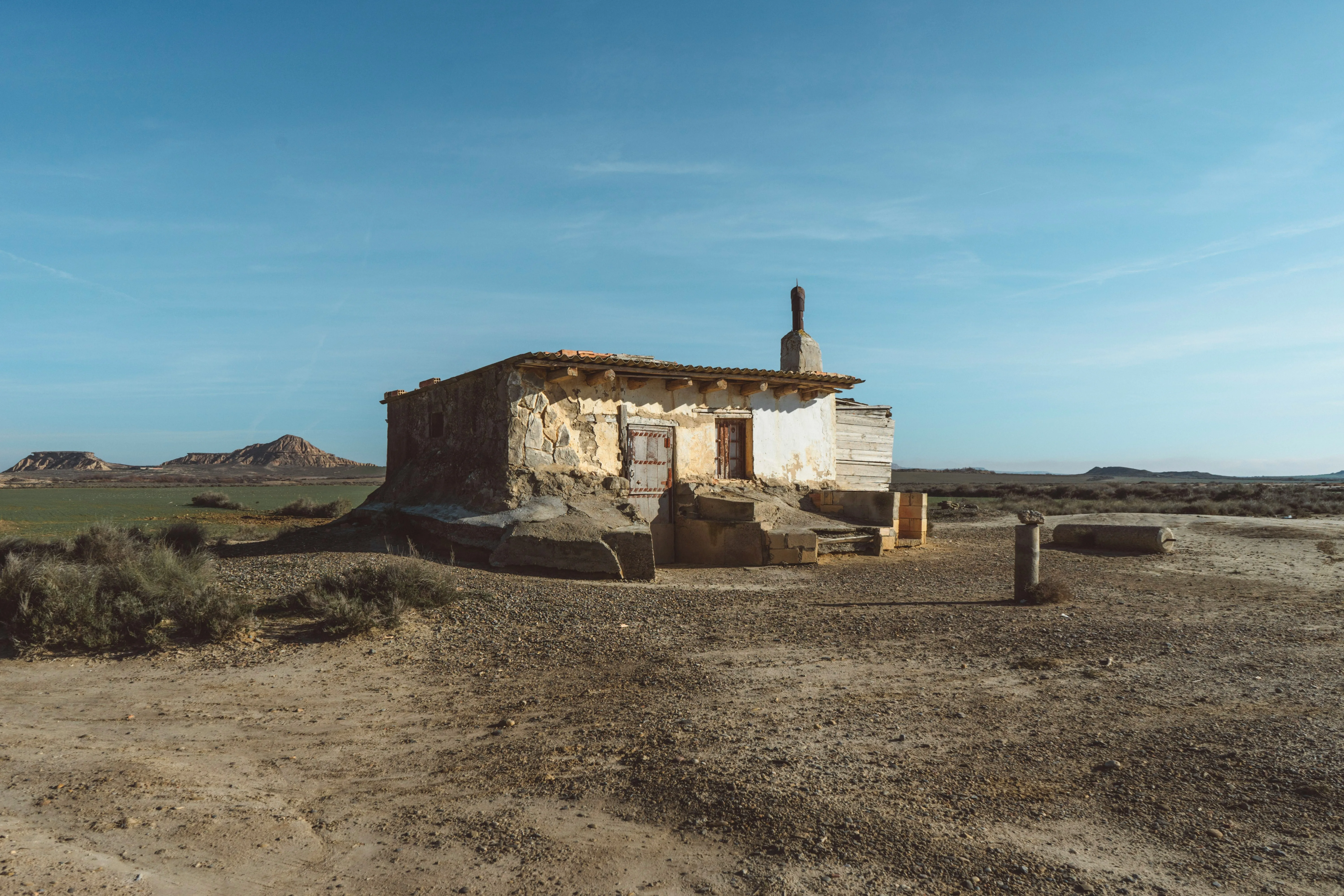 Petit bâtiment en pierre abandonné avec une cheminée, situé dans un paysage semi-désertique aride avec des buttes rocheuses en arrière-plan sous un ciel bleu clair.