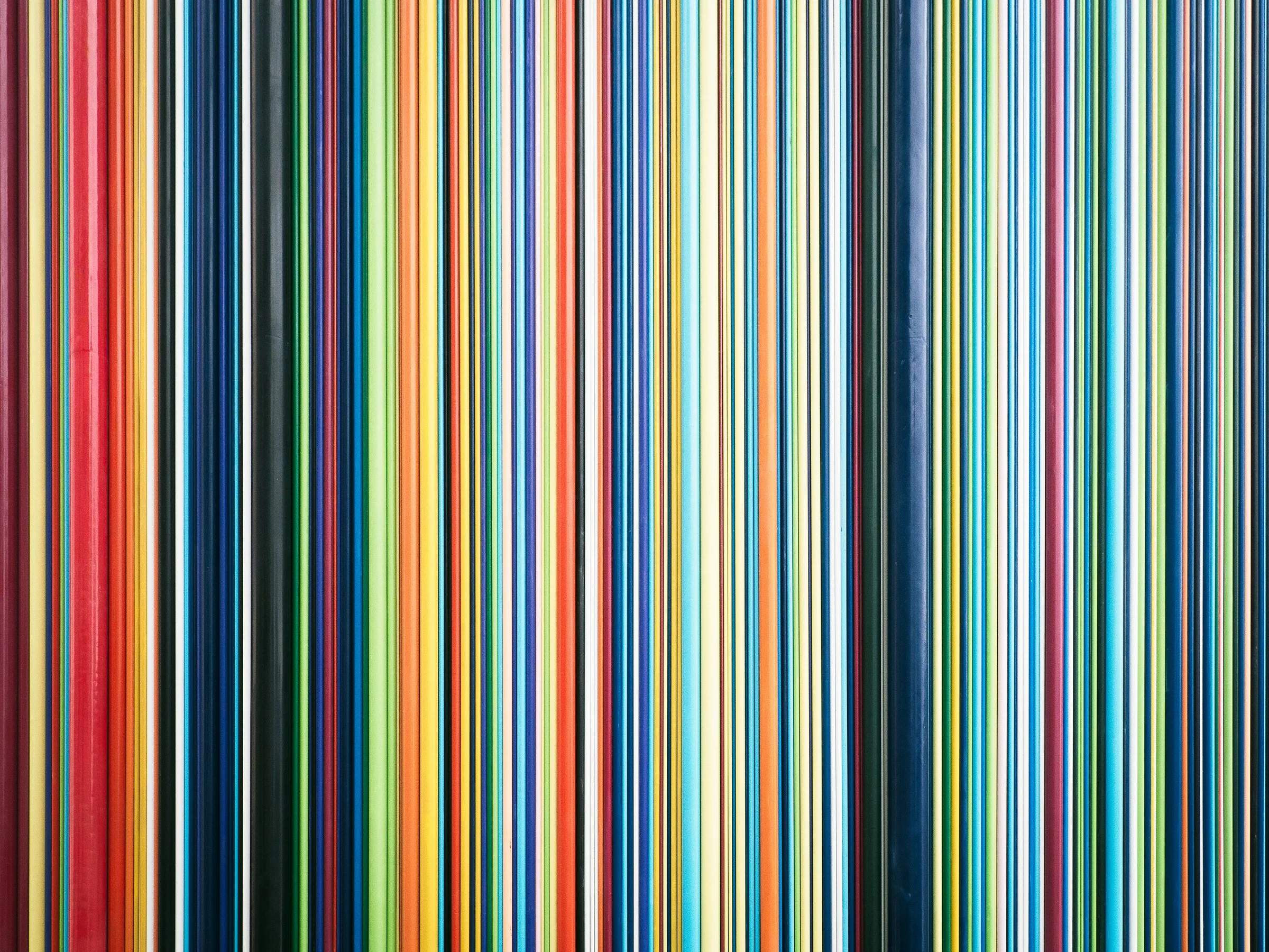 Une surface entièrement couverte de fines bandes verticales aux couleurs vives et variées, incluant rouge, vert, bleu, jaune, orange et blanc, créant un motif graphique abstrait et rythmé.