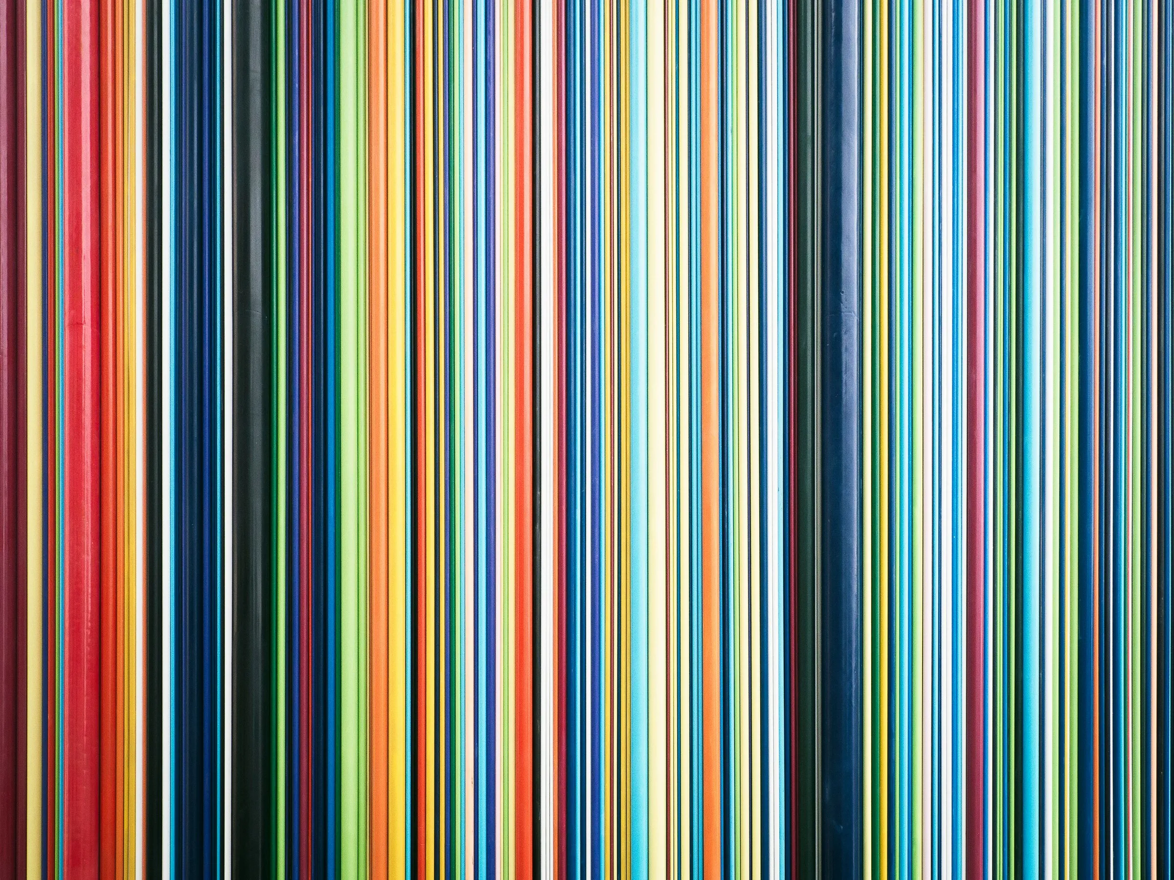 Une surface entièrement couverte de fines bandes verticales aux couleurs vives et variées, incluant rouge, vert, bleu, jaune, orange et blanc, créant un motif graphique abstrait et rythmé.