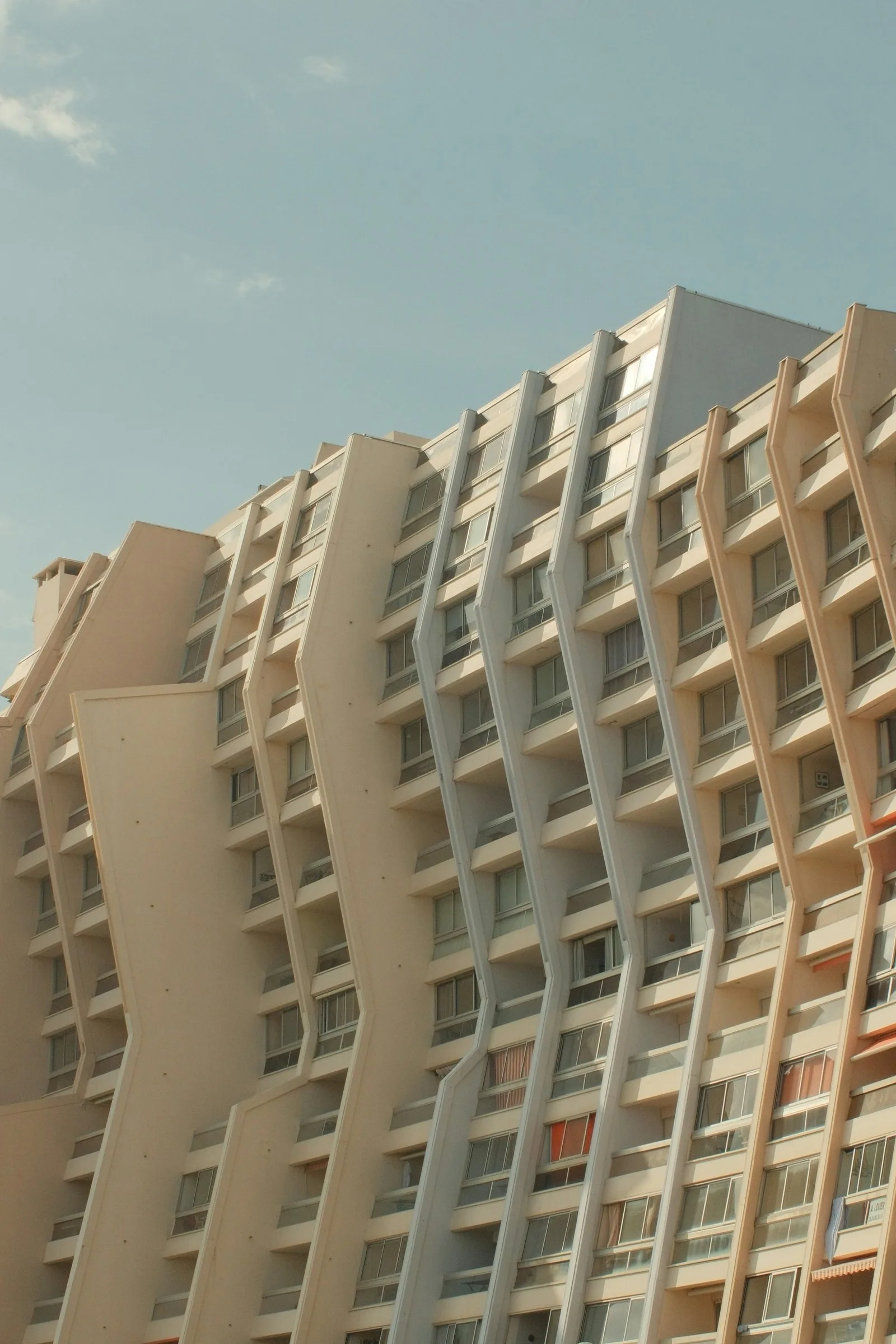 Vue en contre-plongée d'un immeuble résidentiel de grande hauteur avec des nervures verticales en béton beige rythmant la façade, fenêtres et balcons répétitifs sur fond de ciel bleu pâle.