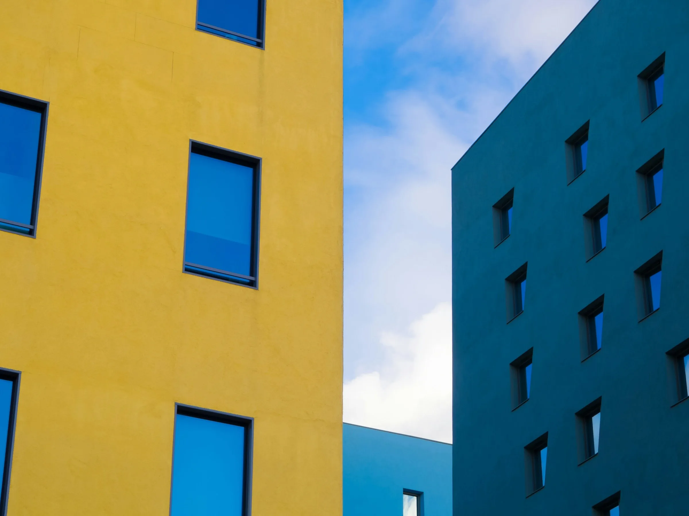 Deux immeubles modernes aux façades contrastées, l'un jaune vif et l'autre bleu-vert, photographiés depuis le bas avec un ciel nuageux visible entre eux.