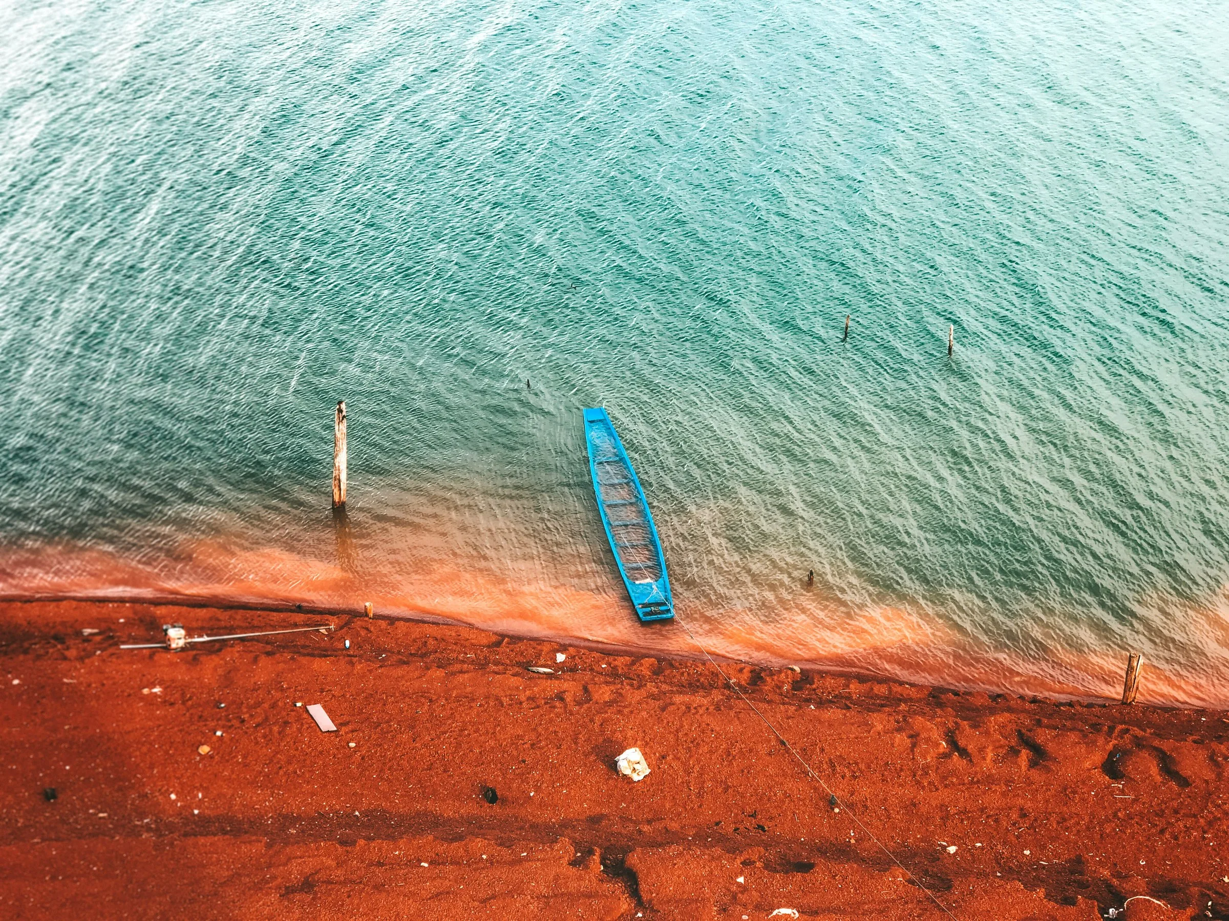 Vue aérienne d'une pirogue bleue posée à la lisière de l'eau turquoise et d'une plage au sable rouge ocre, avec quelques pieux en bois dans l'eau