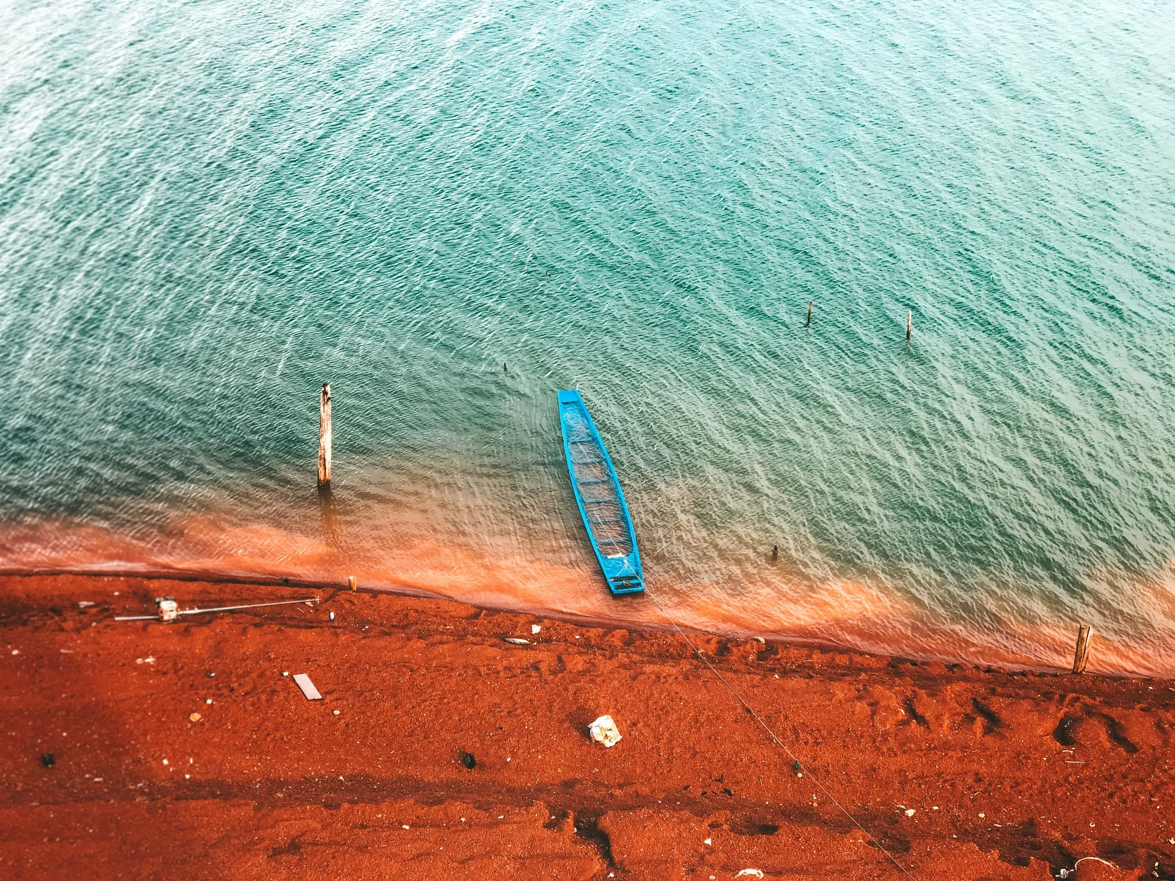 Vue aérienne d'une pirogue bleue posée à la lisière de l'eau turquoise et d'une plage au sable rouge ocre, avec quelques pieux en bois dans l'eau