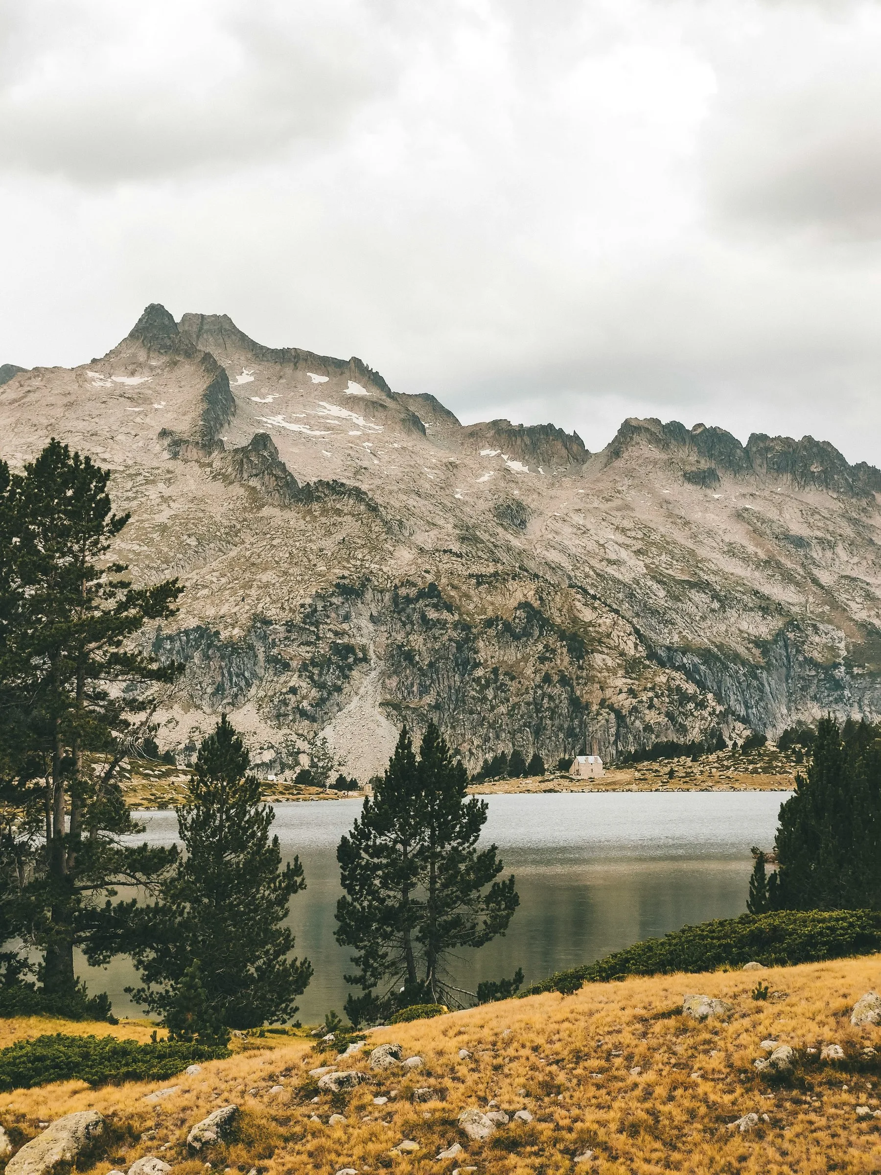Un lac alpin calme entouré de pins et de prairies dorées, avec une petite cabane blanche sur la rive opposée et un massif rocheux imposant en arrière-plan sous un ciel nuageux.