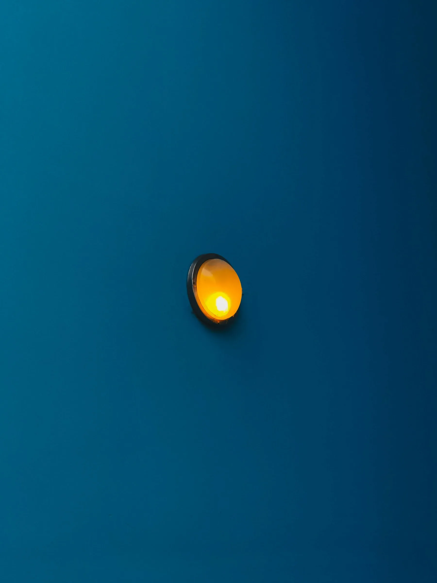 Une applique murale ronde à lumière orange vif montée sur un mur lisse de couleur bleu canard, créant un contraste saisissant entre les deux couleurs complémentaires.