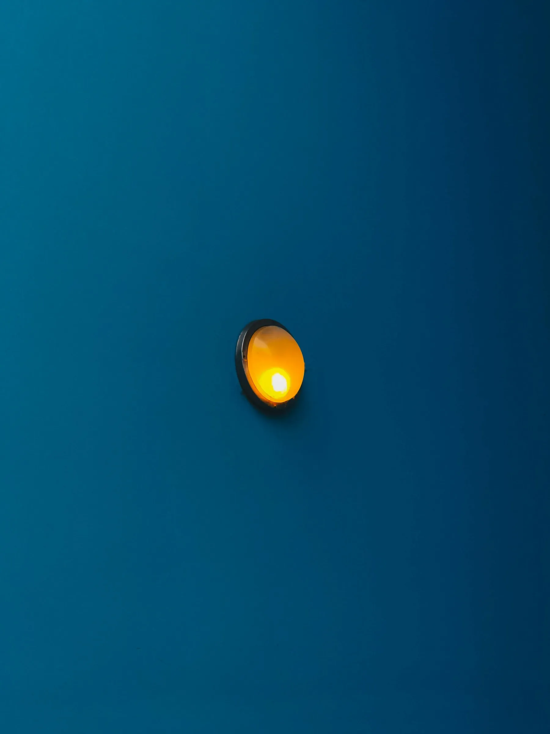 Une applique murale ronde à lumière orange vif montée sur un mur lisse de couleur bleu canard, créant un contraste saisissant entre les deux couleurs complémentaires.