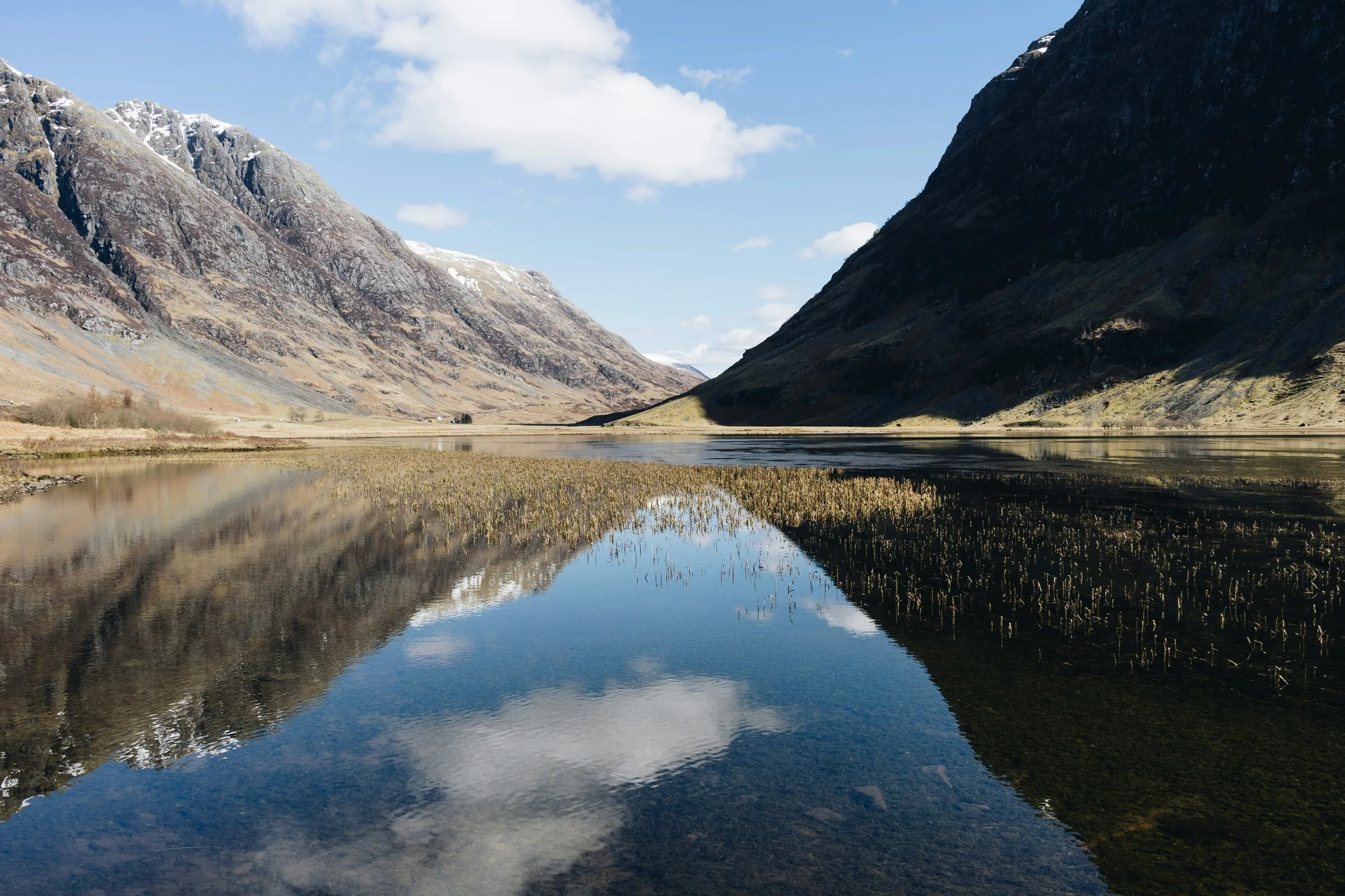 Loch calme reflétant des montagnes enneigées et un ciel bleu nuageux dans une vallée glaciaire écossaise, avec des roseaux émergents au premier plan