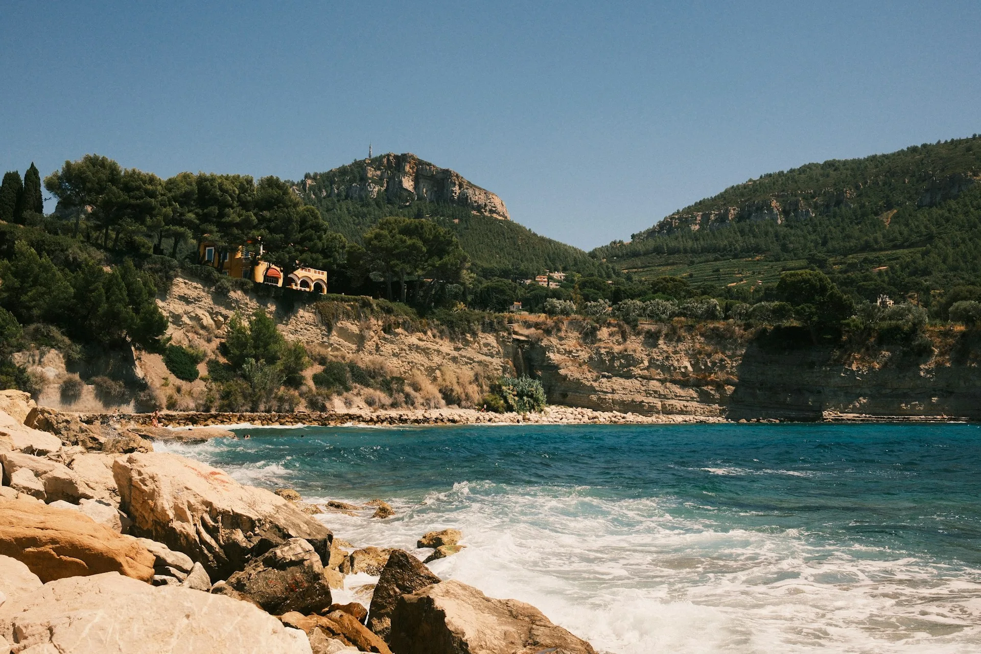 Une crique rocheuse aux eaux turquoise avec des vagues qui s'écrasent sur les rochers au premier plan, bordée de falaises calcaires et de végétation méditerranéenne, avec des maisons perchées sur la colline et des montagnes boisées en arrière-plan.