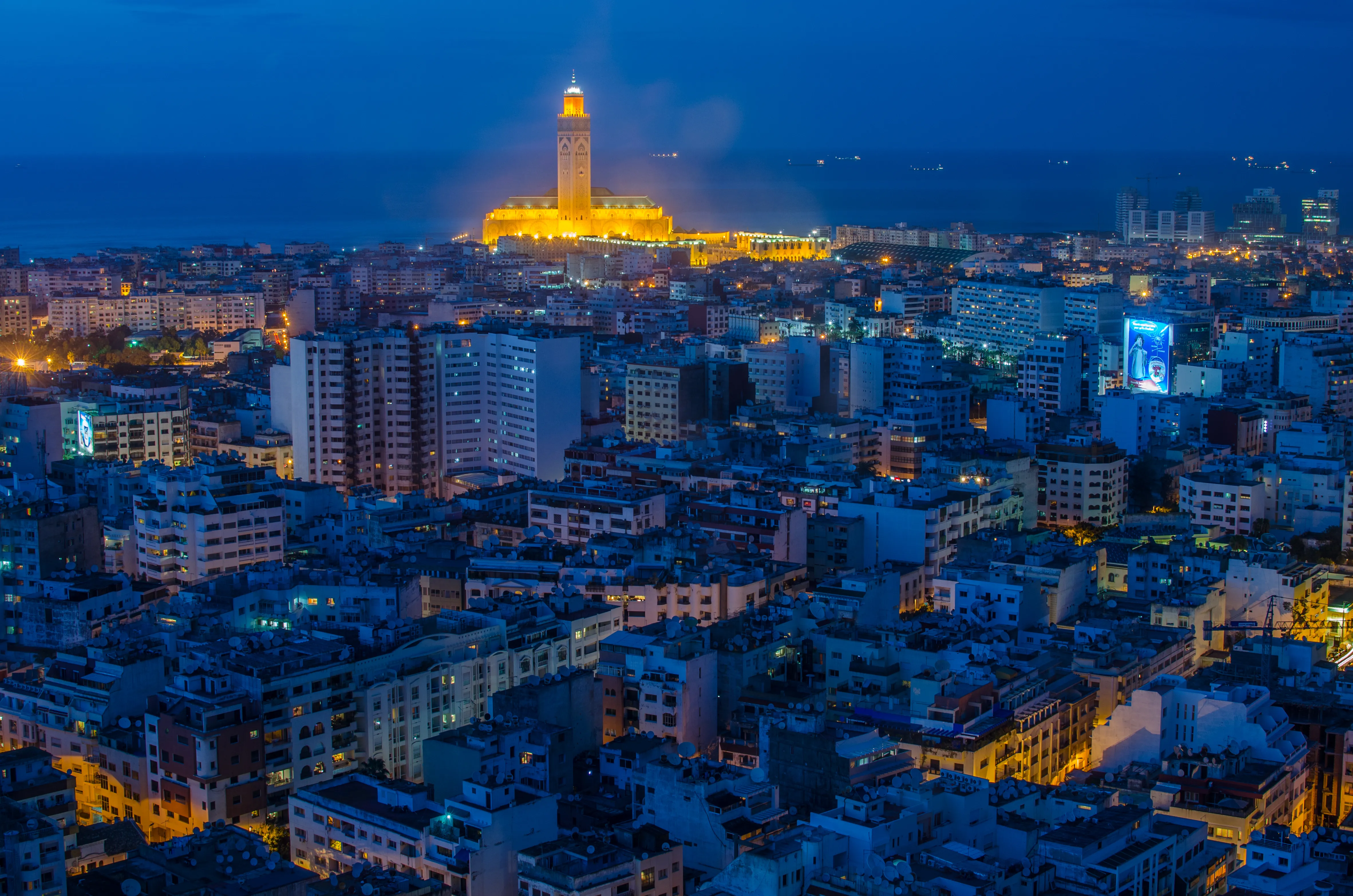 Vue aérienne nocturne de Casablanca avec la mosquée Hassan II illuminée en orange au bord de l'océan Atlantique, dominant un vaste tissu urbain baigné de lumières bleues.