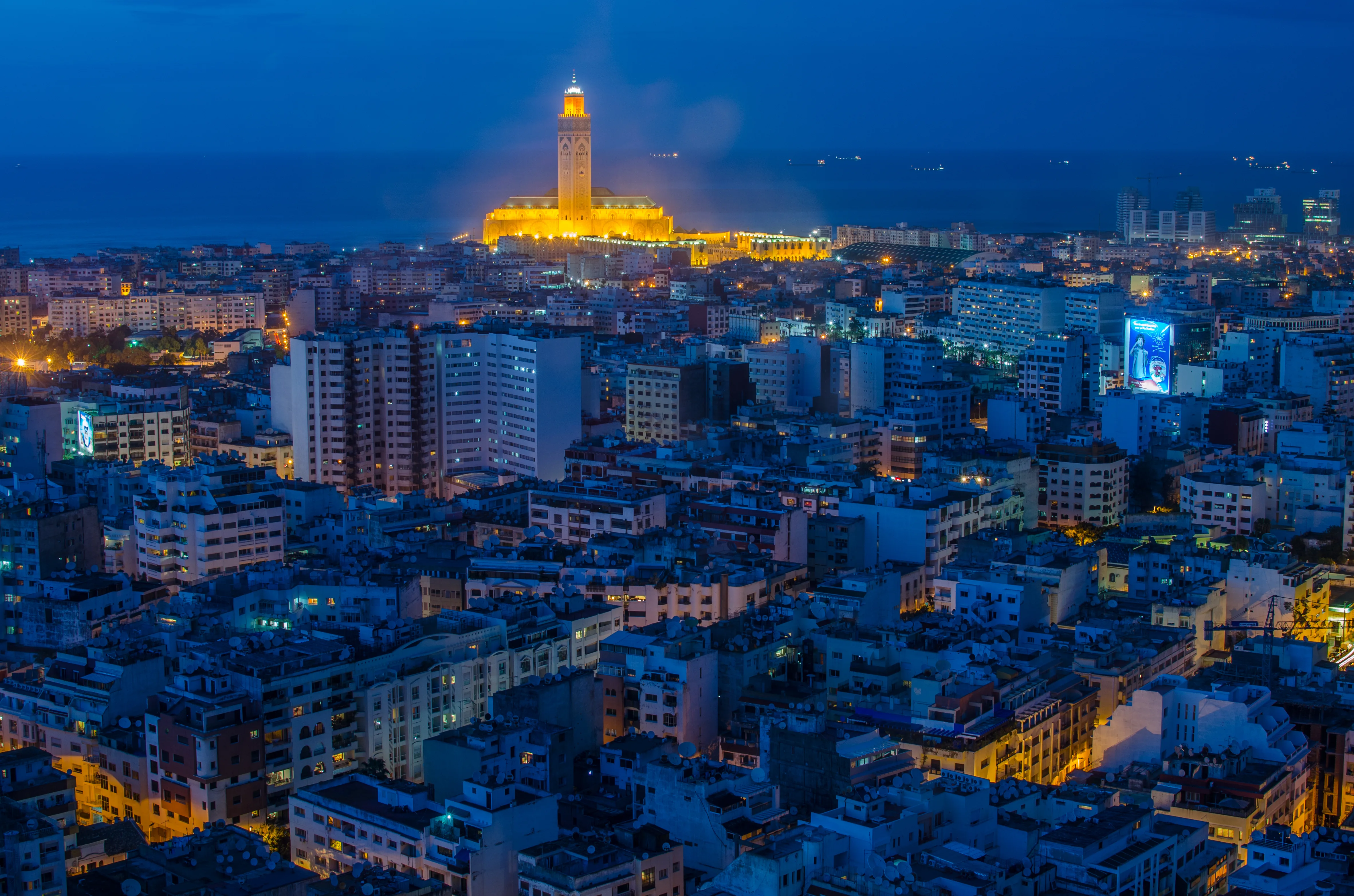 Vue aérienne nocturne de Casablanca avec la mosquée Hassan II illuminée en orange au bord de l'océan Atlantique, dominant un vaste tissu urbain baigné de lumières bleues.