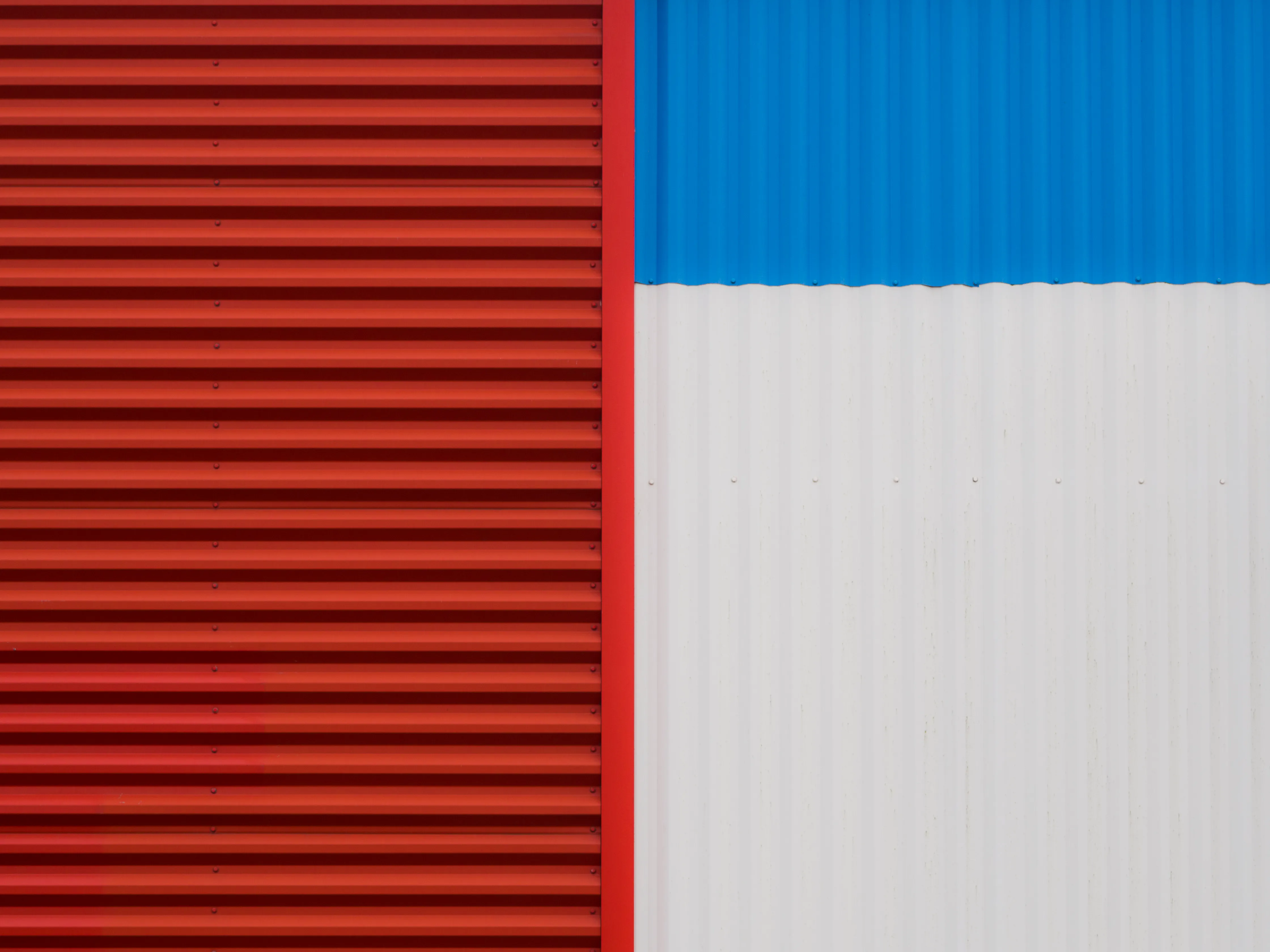Mur en tôle ondulée divisé en deux sections : à gauche un pan entièrement rouge, à droite une partie blanche surmontée d'un bandeau bleu, formant une composition géométrique abstraite.