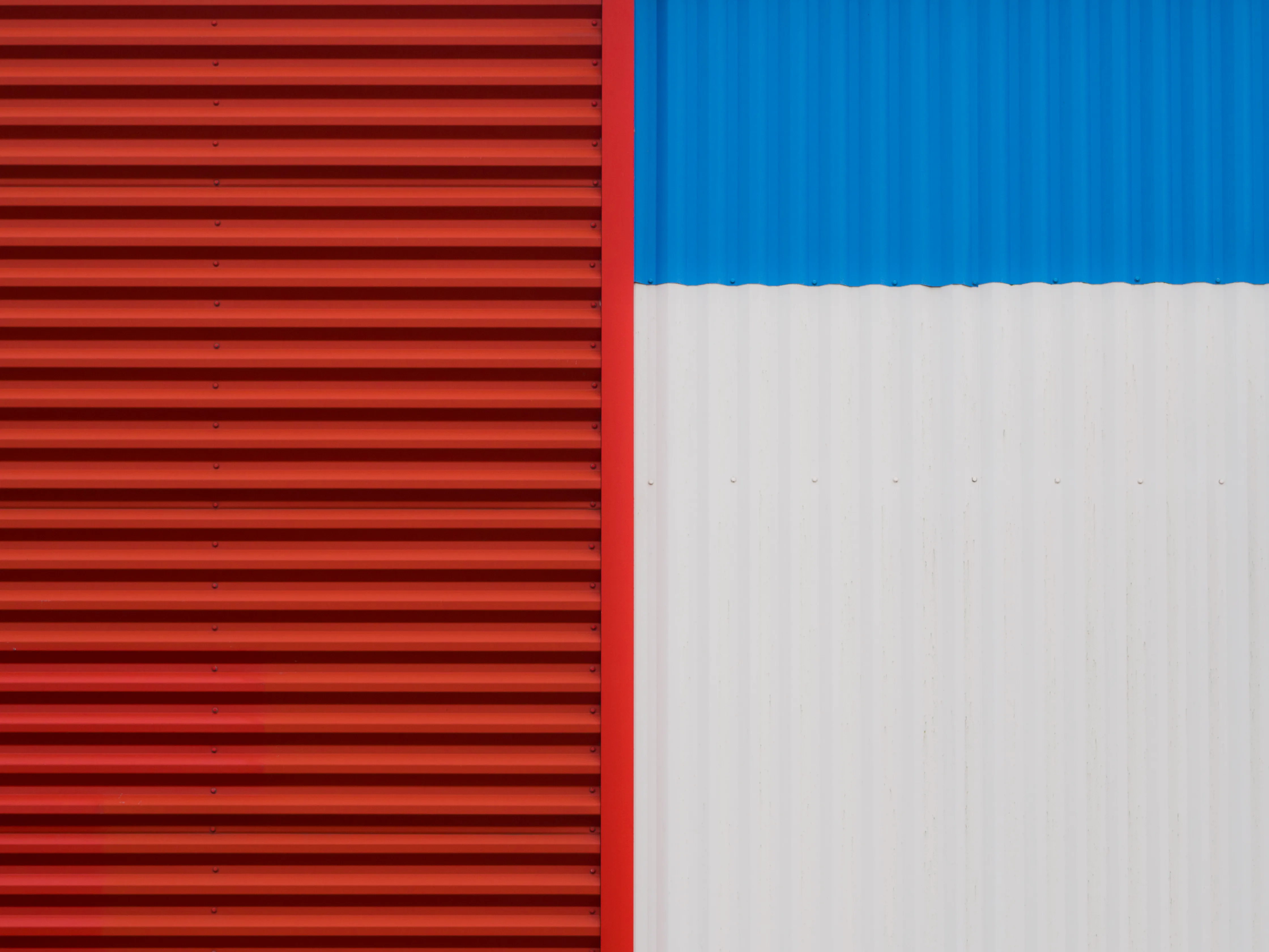 Mur en tôle ondulée divisé en deux sections : à gauche un pan entièrement rouge, à droite une partie blanche surmontée d'un bandeau bleu, formant une composition géométrique abstraite.