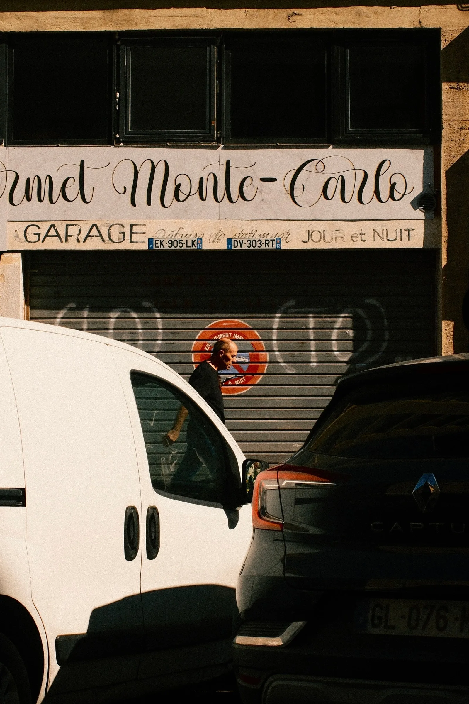 Façade d'un garage ancien portant l'enseigne Monte-Carlo avec un homme passant entre une camionnette blanche et un SUV Renault Captur, baignée d'une lumière dorée rasante