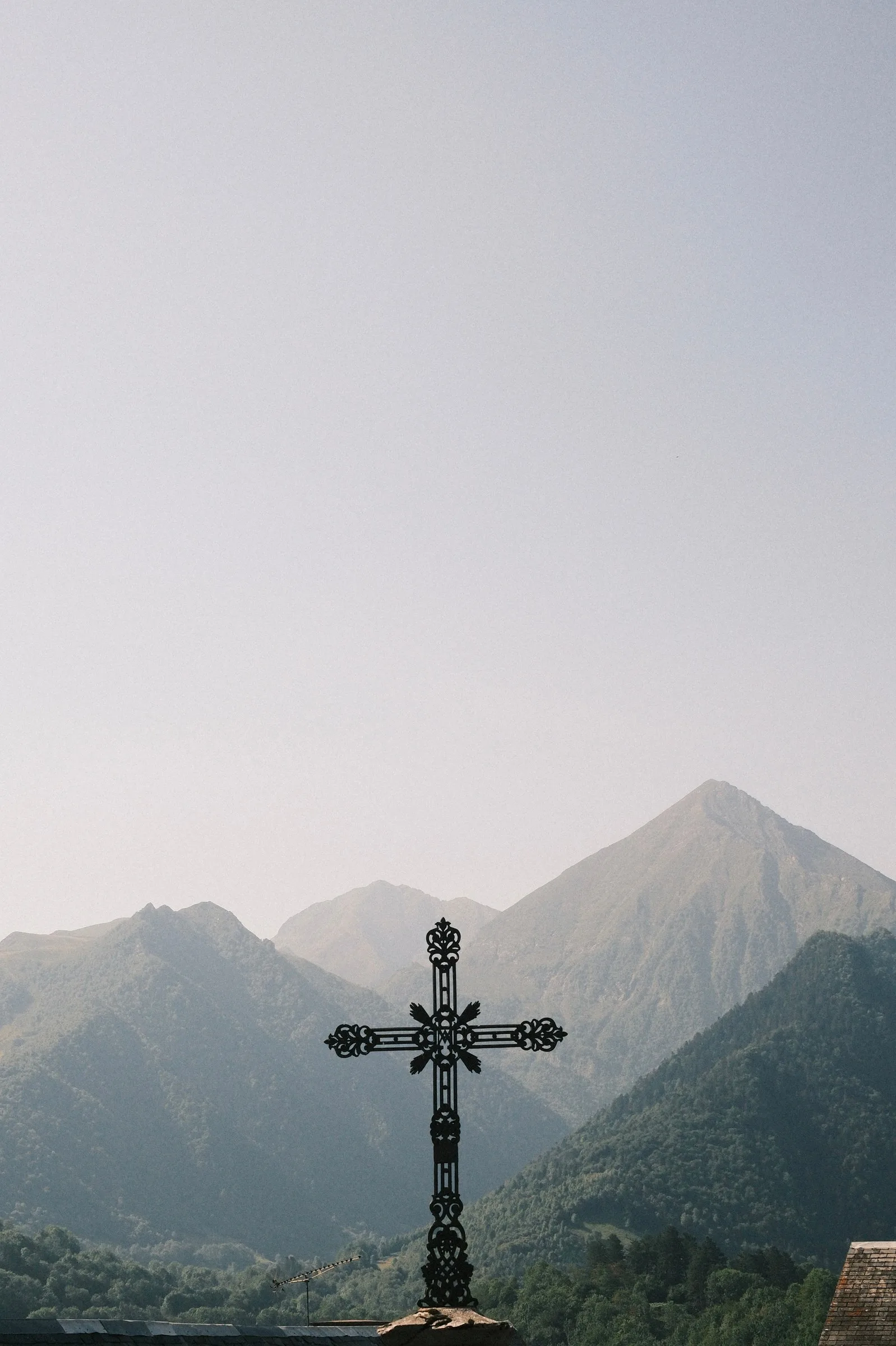 Une croix ornementale en fer forgé noir se détache devant un massif montagneux enveloppé de brume, sous un ciel clair et lumineux.
