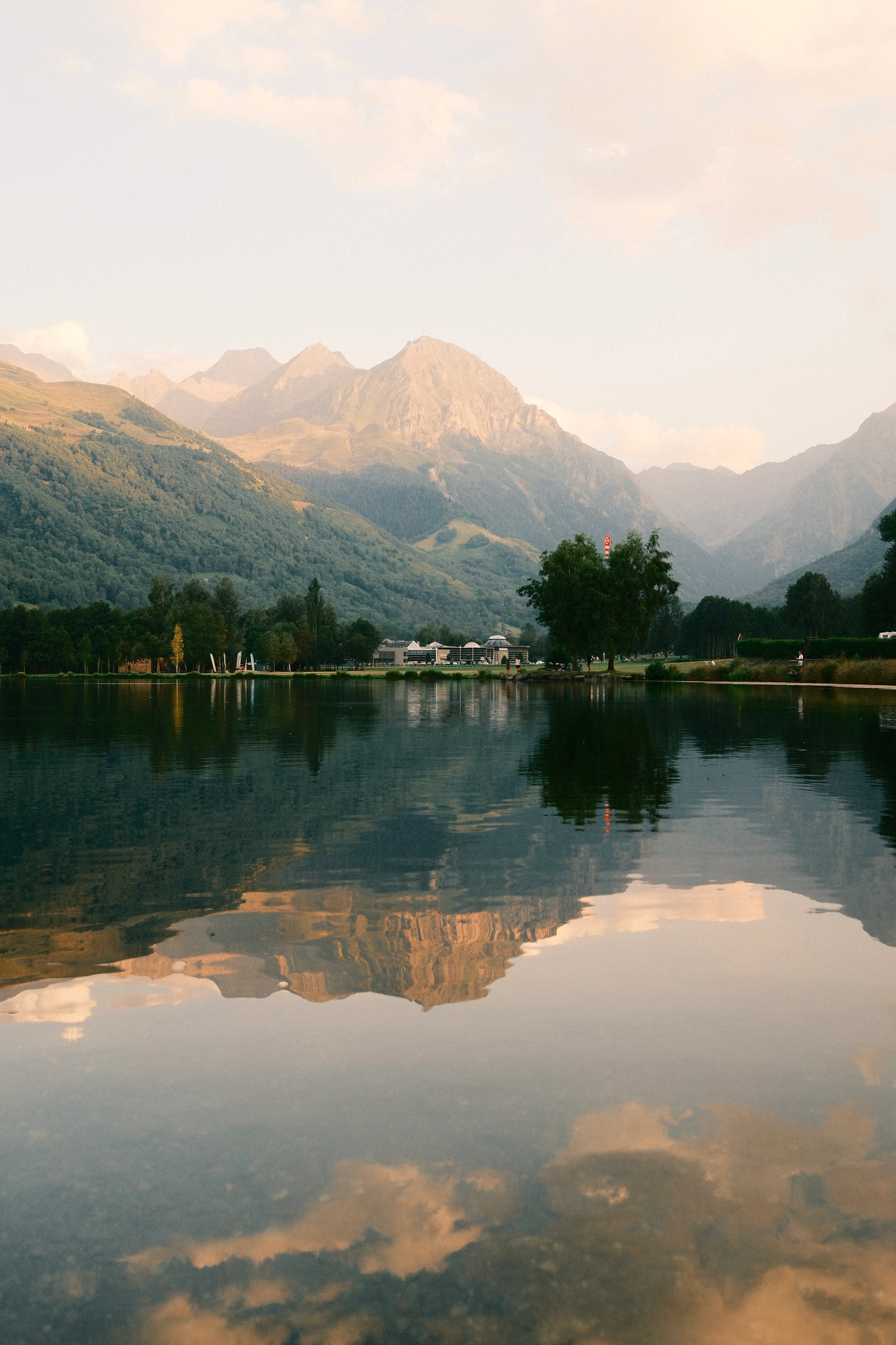 Lac de montagne aux eaux calmes reflétant des sommets alpins et des nuages dorés, avec une bâtisse et des arbres sur la rive opposée, au coucher du soleil.