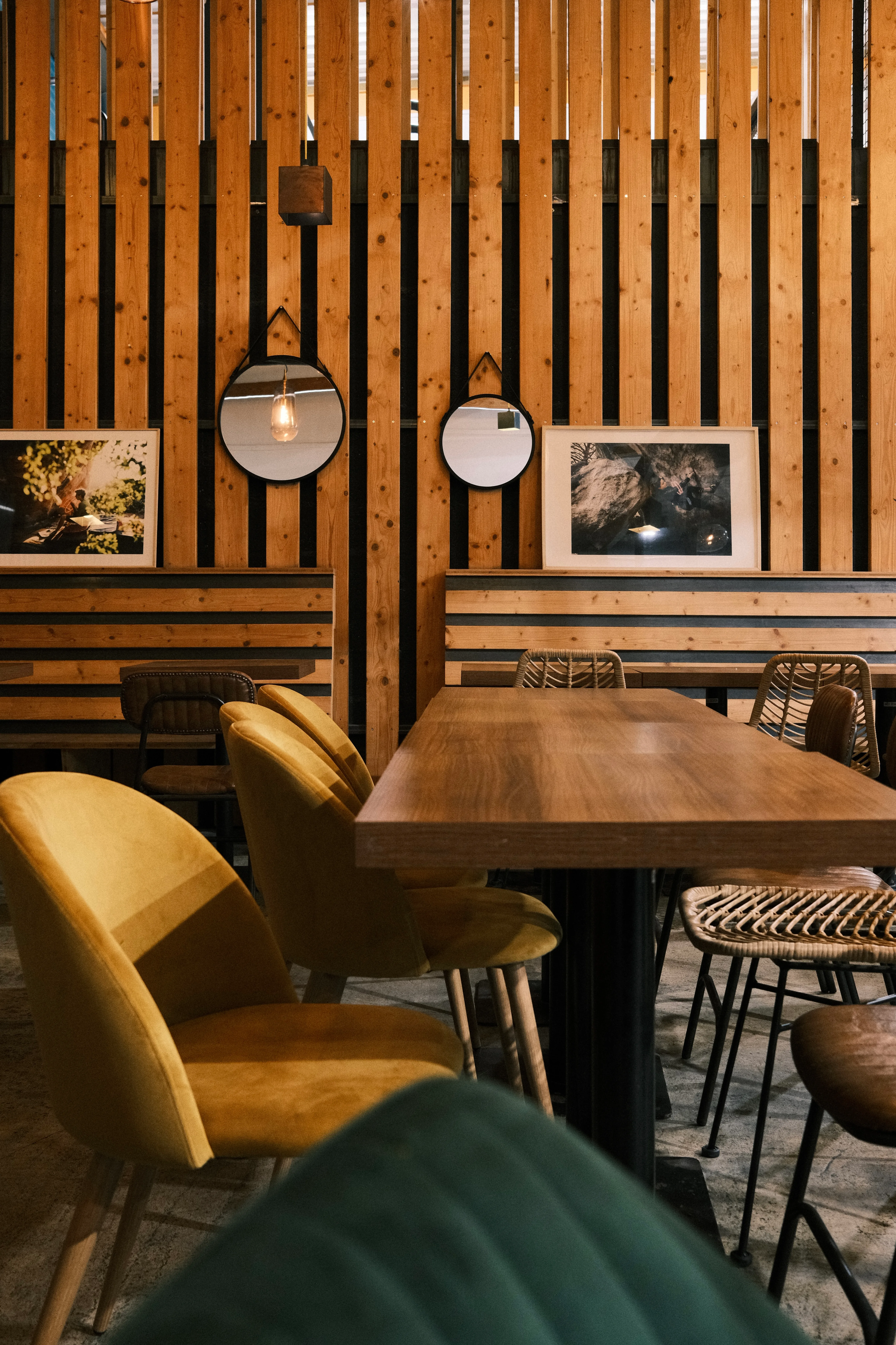 Intérieur d'un restaurant ou café avec des chaises en velours jaune moutarde, une table en bois massif, des miroirs ronds et des lambris en bois verticaux ornés de cadres photographiques.