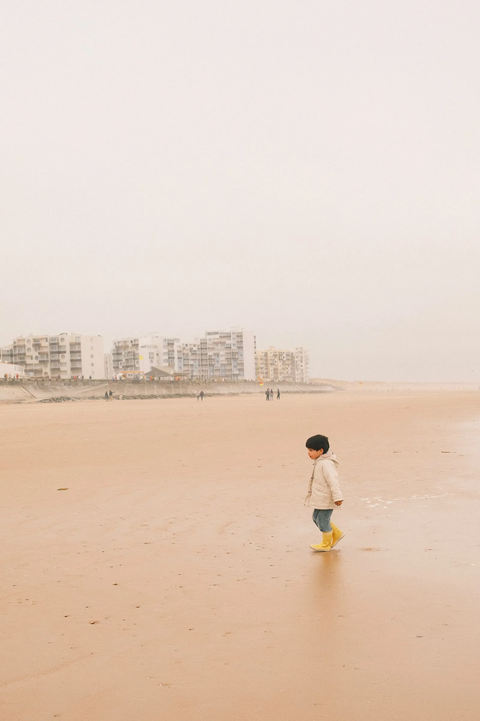 Un jeune enfant vêtu d'un manteau beige et de bottes en caoutchouc jaunes marche seul sur une vaste plage de sable mouillé par temps brumeux, avec des immeubles côtiers en arrière-plan.