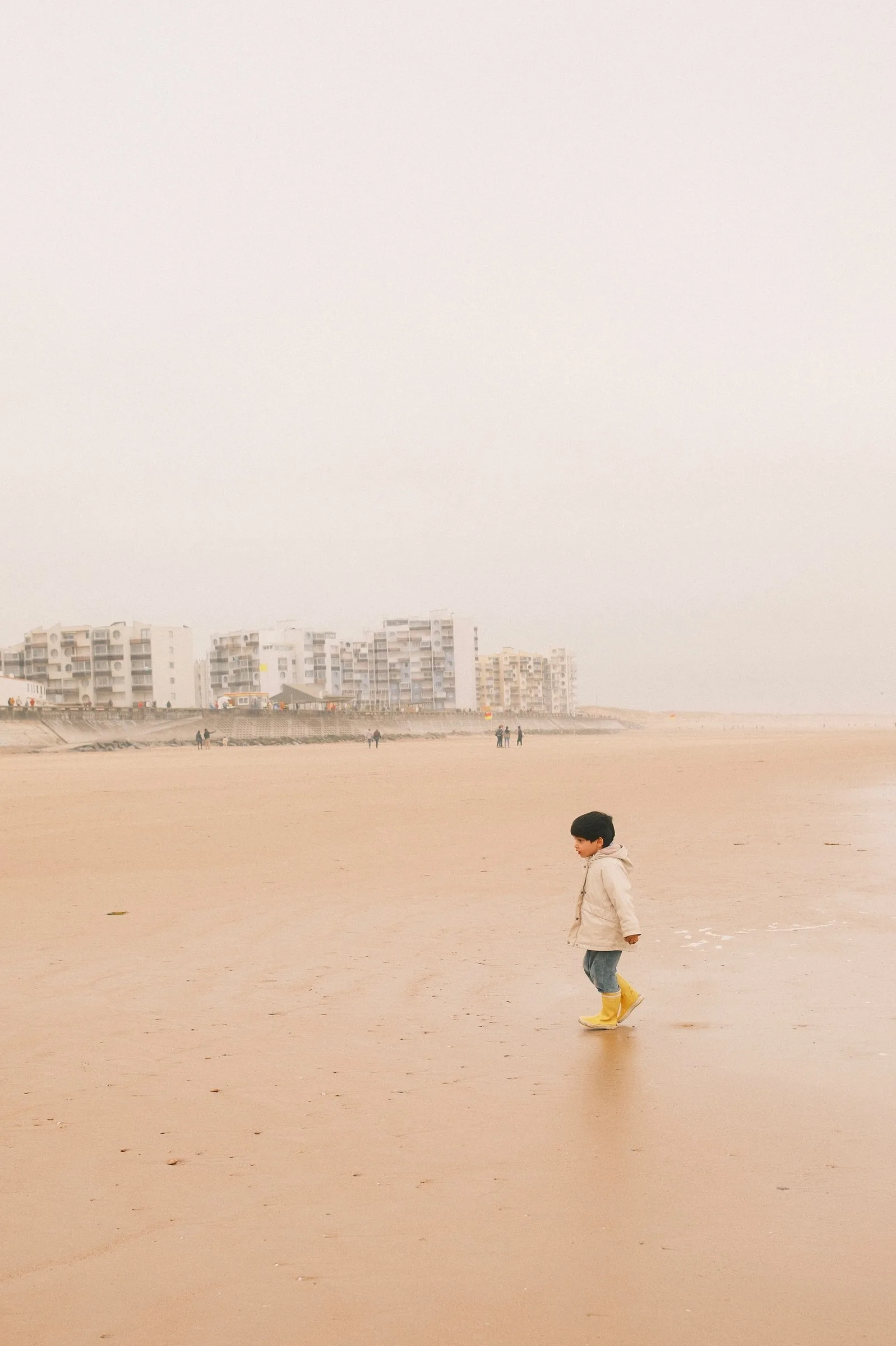 Un jeune enfant vêtu d'un manteau beige et de bottes en caoutchouc jaunes marche seul sur une vaste plage de sable mouillé par temps brumeux, avec des immeubles côtiers en arrière-plan.