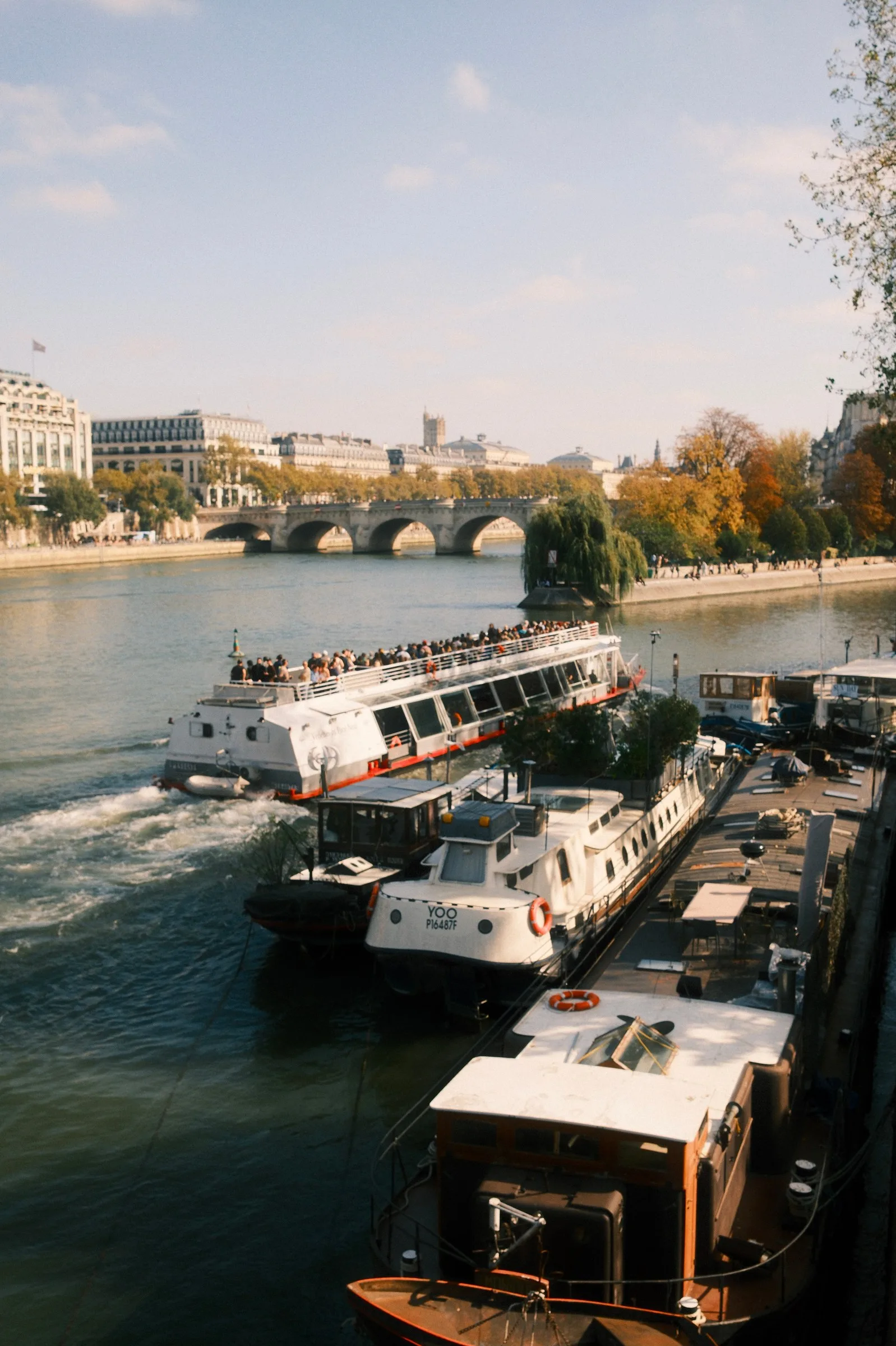 Vue en hauteur de bateaux touristiques et péniches amarrés sur la Seine à Paris, avec le Pont Neuf et les bâtiments haussmanniens en arrière-plan sous un ciel légèrement nuageux.