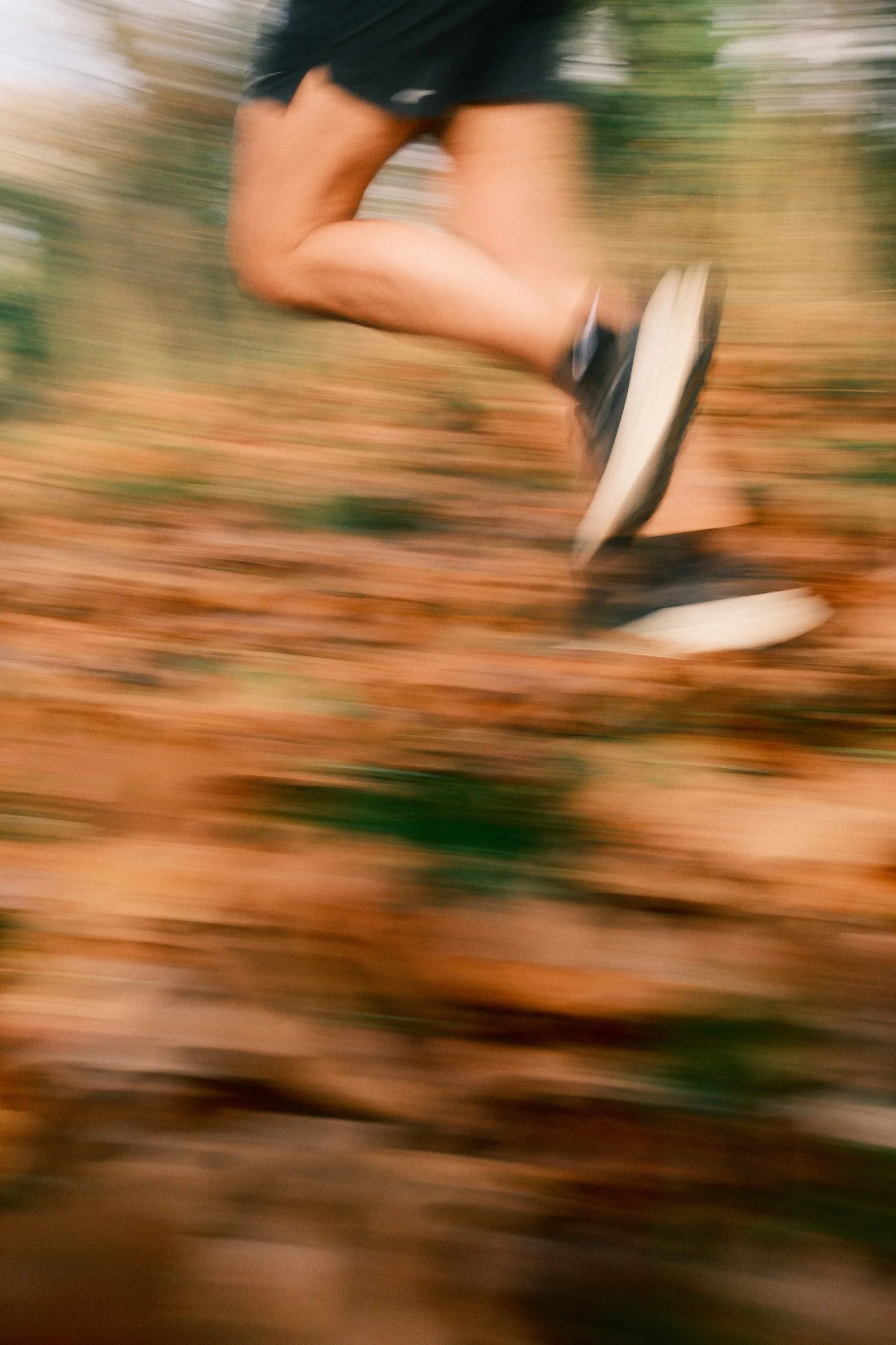 Gros plan flou sur les jambes d'un coureur portant des baskets noires et blanches, en pleine course sur un sol recouvert de feuilles mortes brunes en forêt.