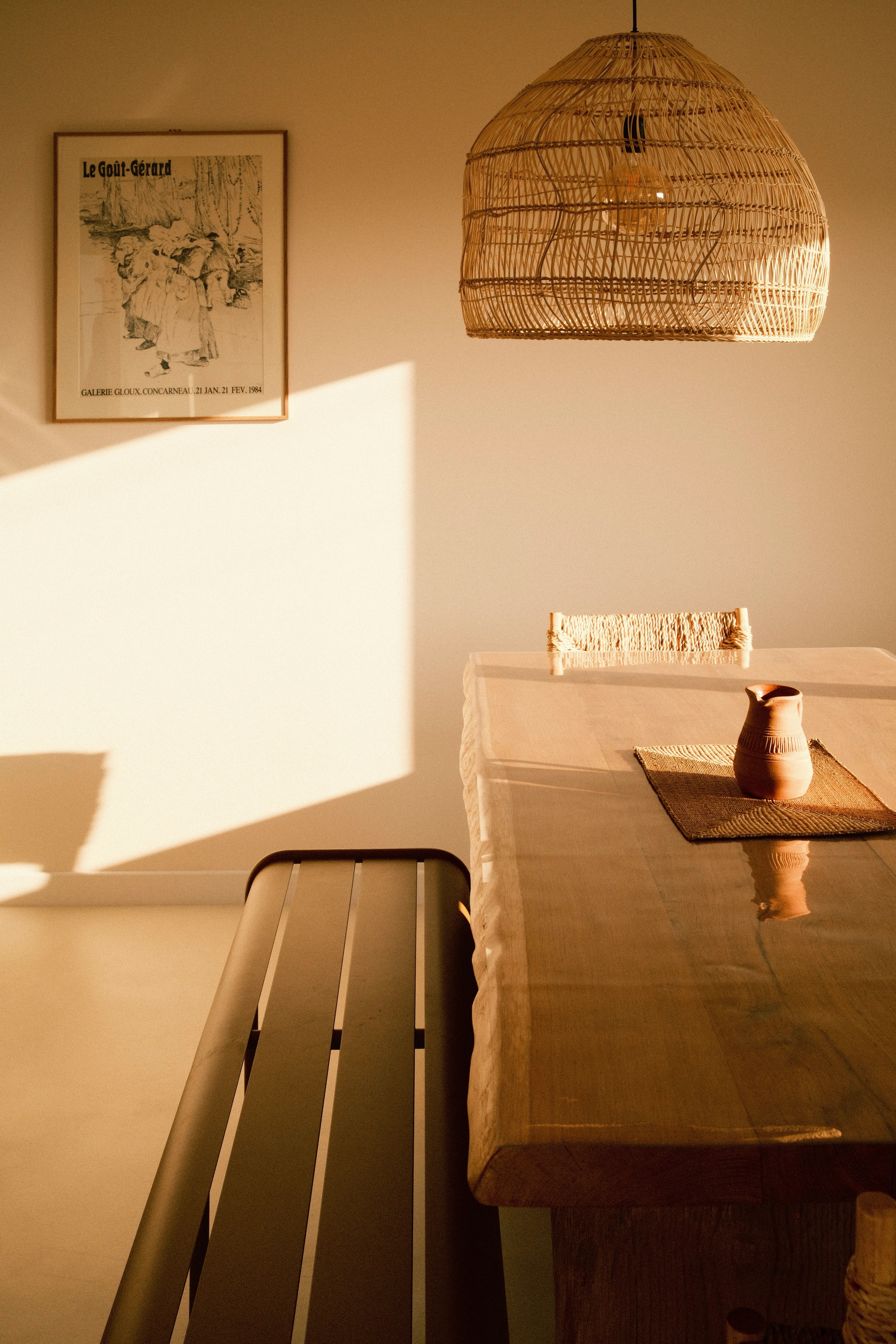 Table en bois recouverte d'un linge blanc avec une cruche en terre cuite, éclairée par une suspension en rotin tressé et la lumière chaude du soleil couchant. Une affiche encadrée 'Le Goût-Gérard' est accrochée au mur.