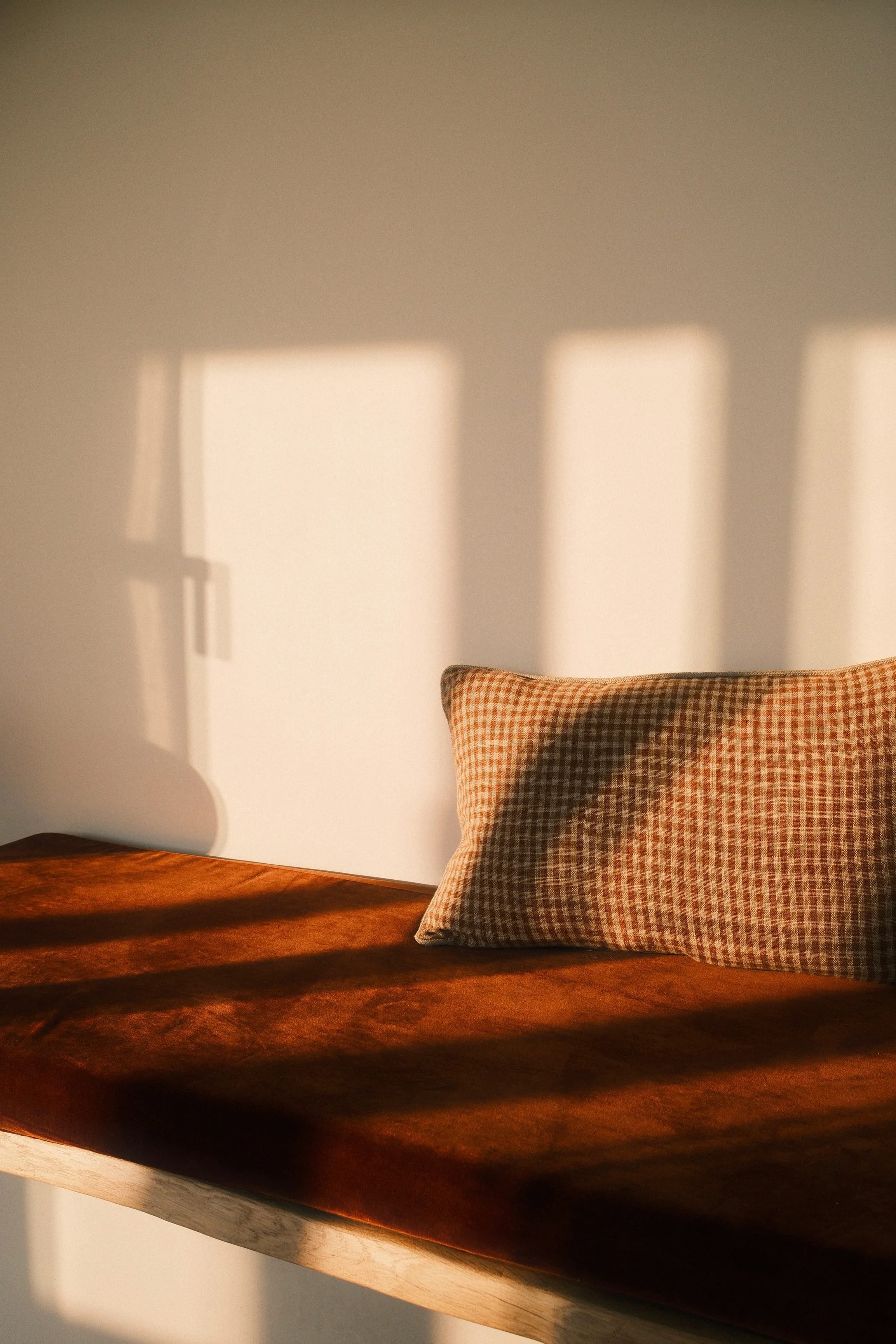 Un coussin à motif vichy brun et beige posé sur un banc en bois massif teinté acajou, éclairé par une lumière naturelle dorée projetant des ombres géométriques sur un mur blanc.