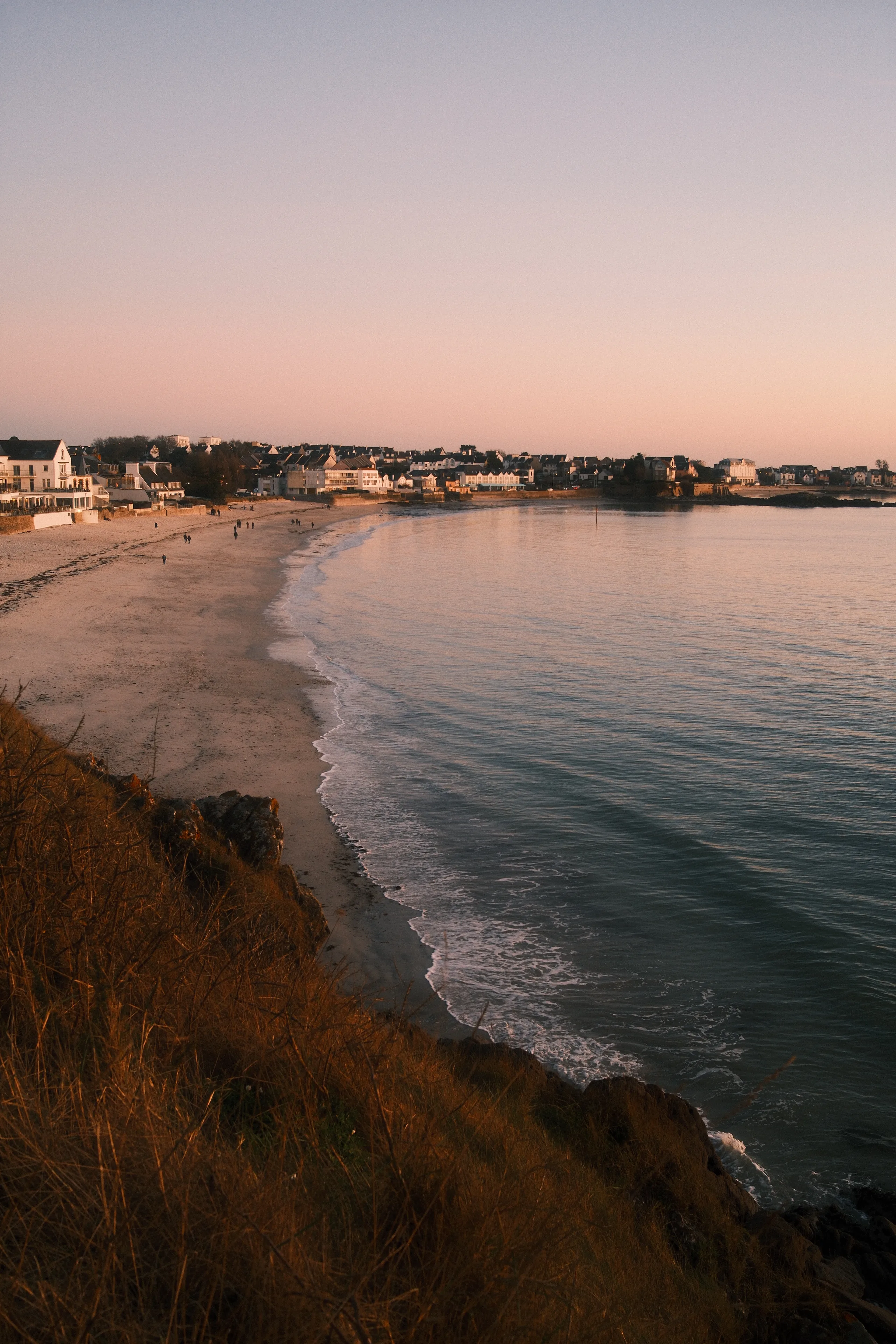 Vue panoramique depuis une falaise rocheuse sur une plage en arc de cercle baignée d'une lumière dorée au coucher du soleil, avec un village côtier en arrière-plan et des promeneurs sur le sable.