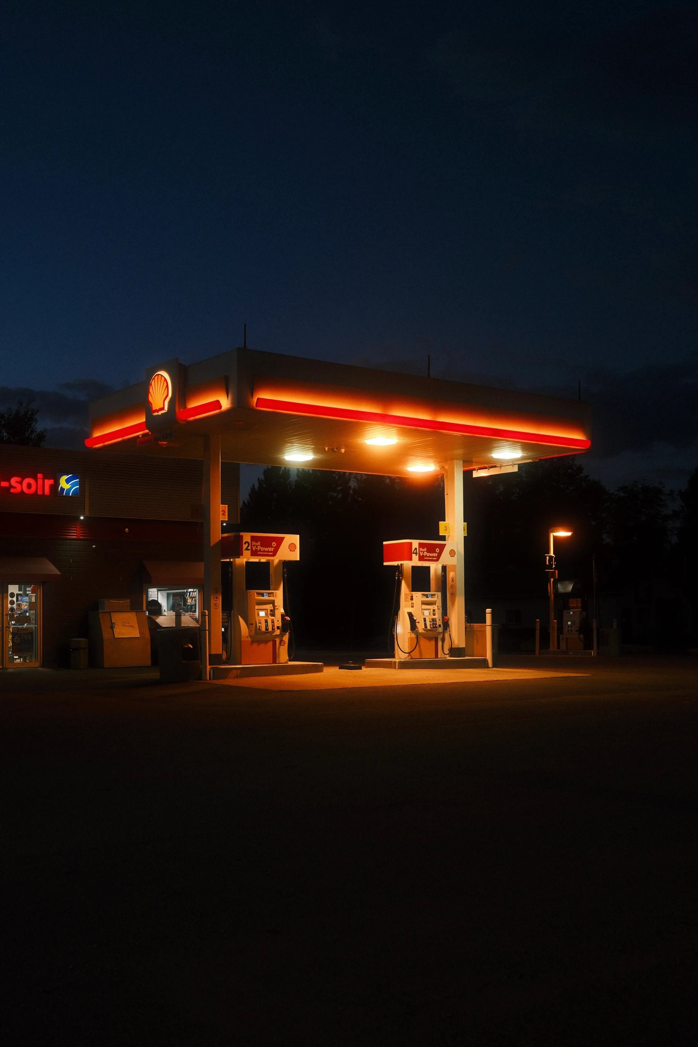 Une station-service Shell déserte photographiée de nuit, avec son auvent lumineux aux néons rouges et orangés éclairant deux pompes à carburant, sur fond de ciel bleu noctambule et d'arbres sombres.