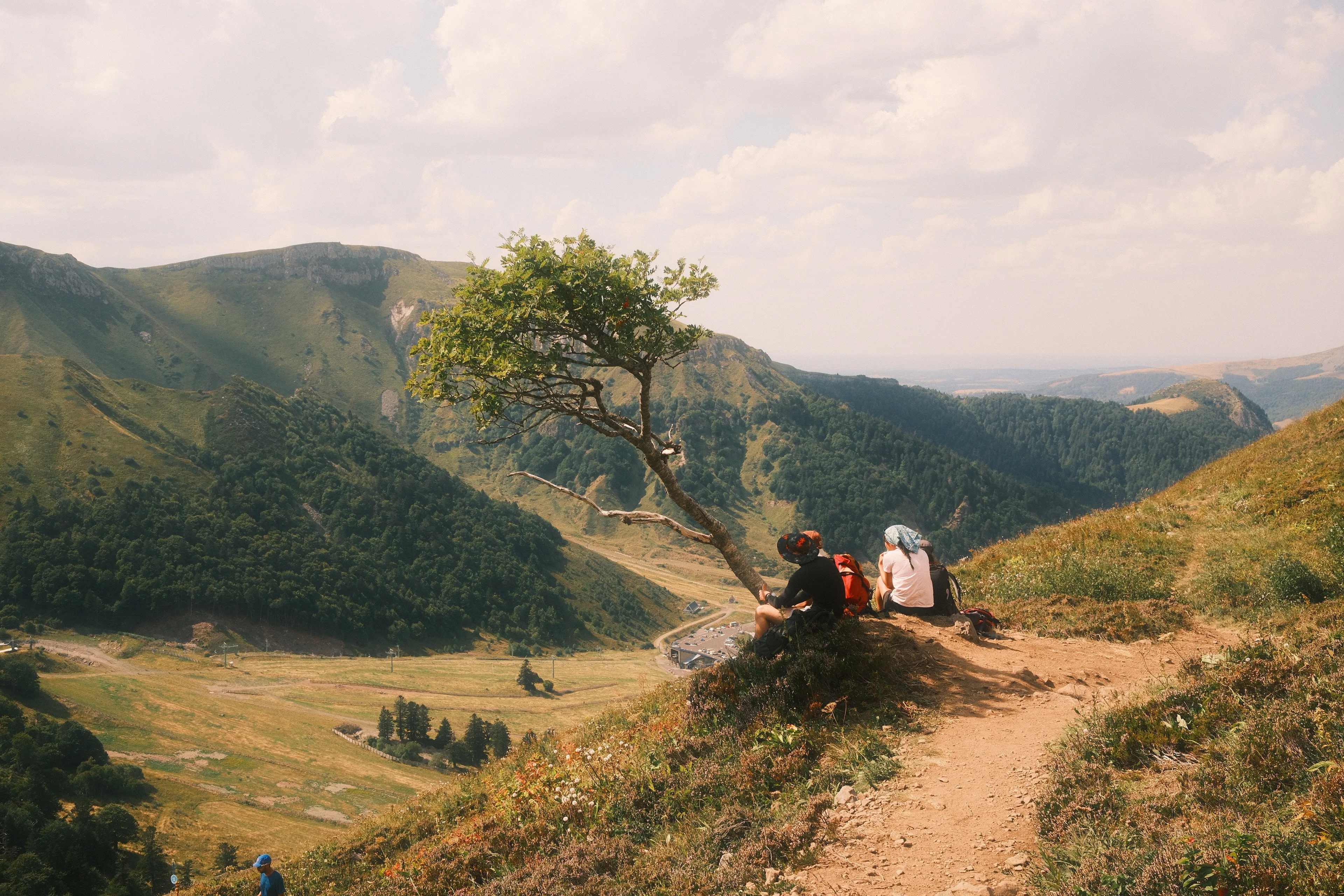 Deux randonneurs assis sous un arbre isolé sur un sentier de montagne, contemplant un vaste paysage de vallées verdoyantes et de sommets sous un ciel nuageux.