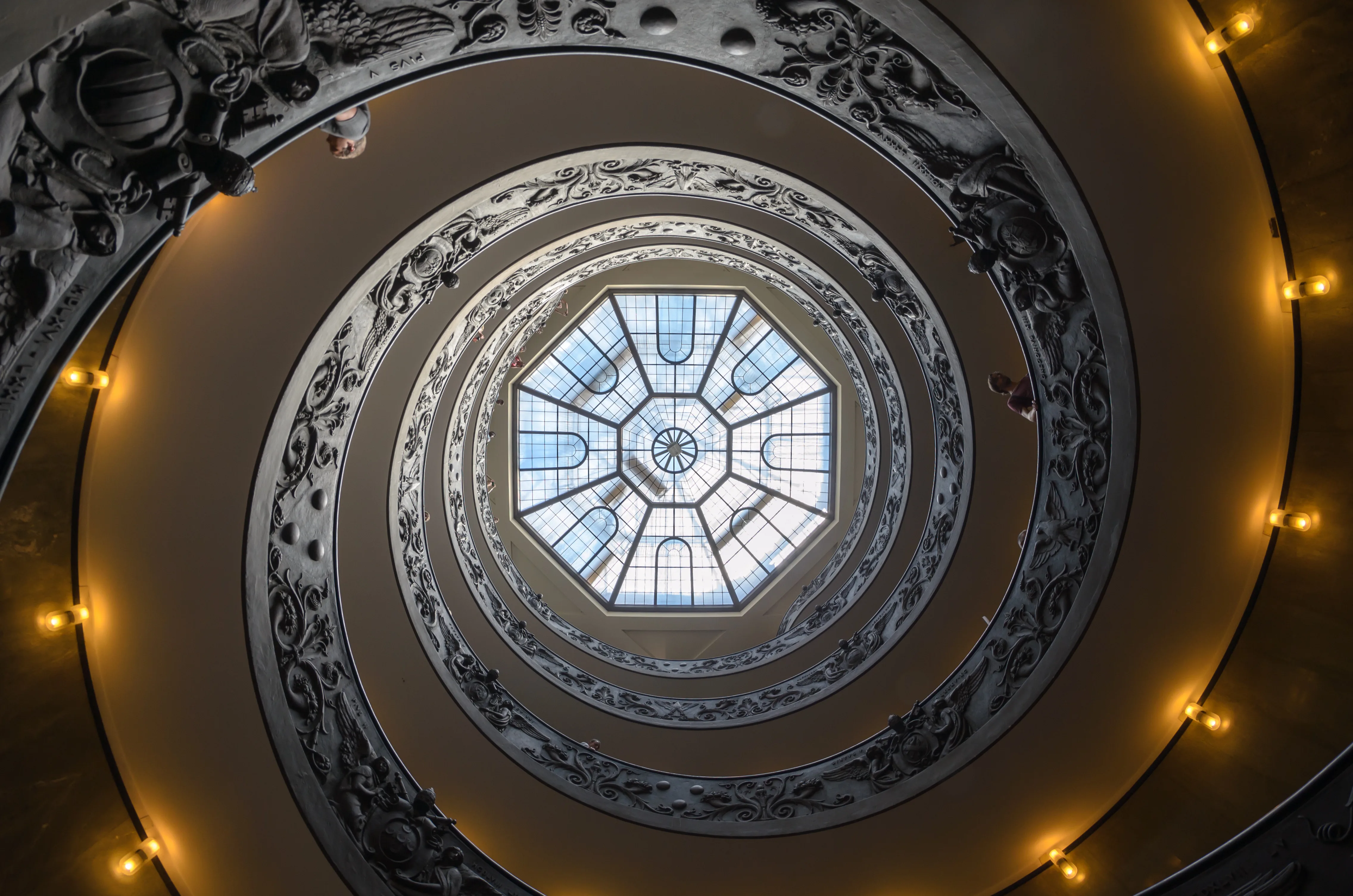 Vue en plongée vers le haut d'un escalier hélicoïdal orné de sculptures baroques, avec une verrière octogonale lumineuse en son centre et des appliques lumineuses dorées le long de la rampe